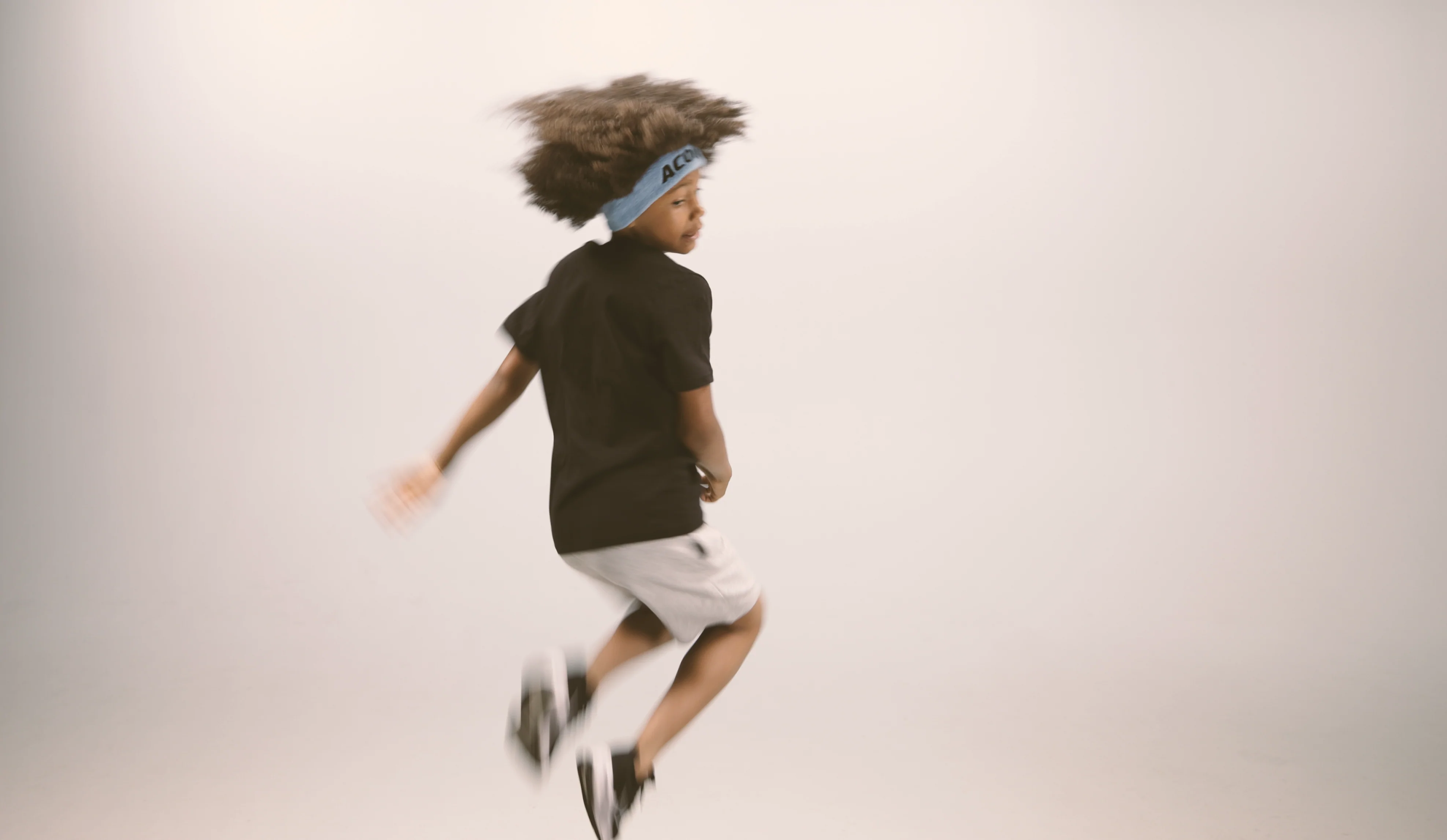 A jumping kid wearing an Acon headband, black t-shirt, white shorts and sneakers. Image is taken against beige background.