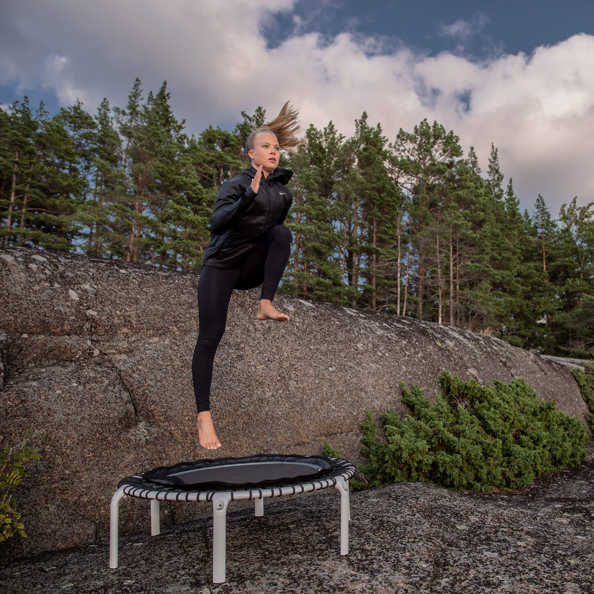 A woman is jumping on a white Acon FIT trampoline in a Nordic forest landscape.