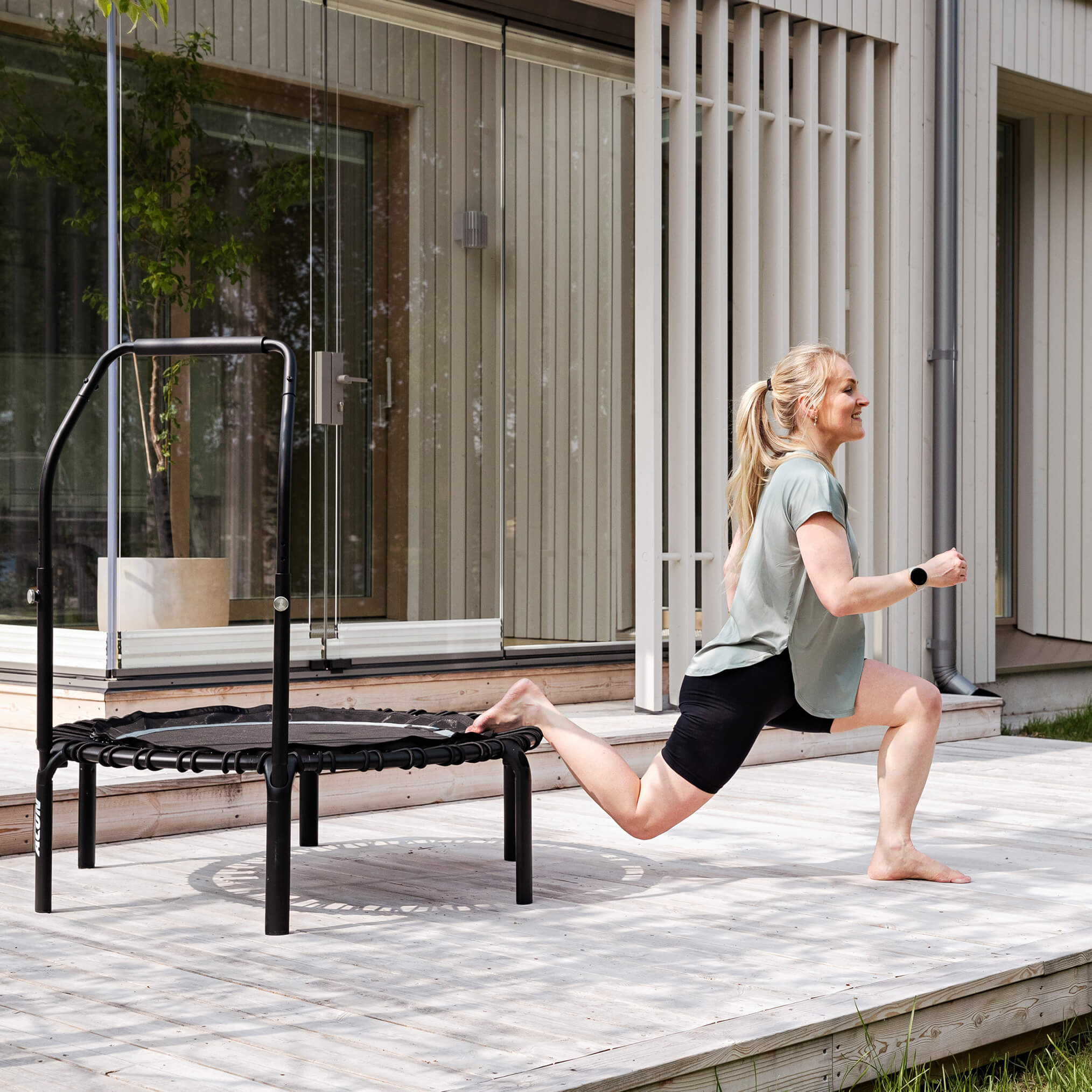 A woman is doing single-leg squats on a black Acon FIT trampoline on a terrace.