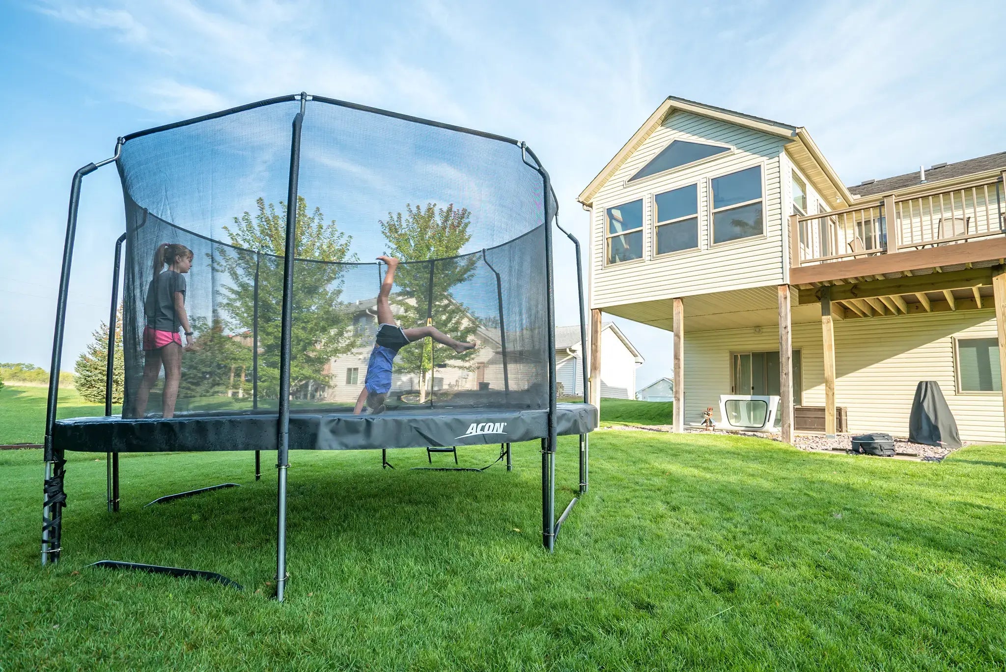 Two kids jumping on an Acon trampoline, one is doing a trick while other one is watching.