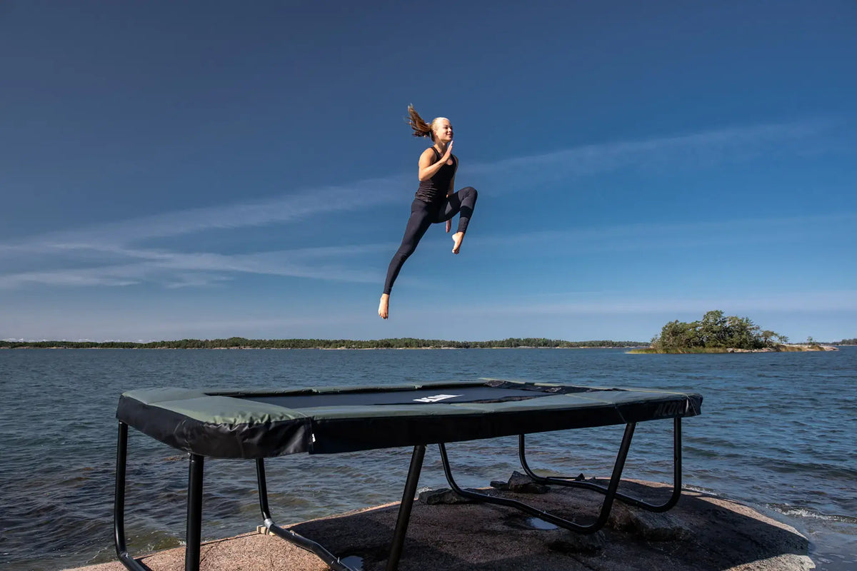 Person jumping high on a rectangular Acon trampoline set on a rocky shore by the sea.