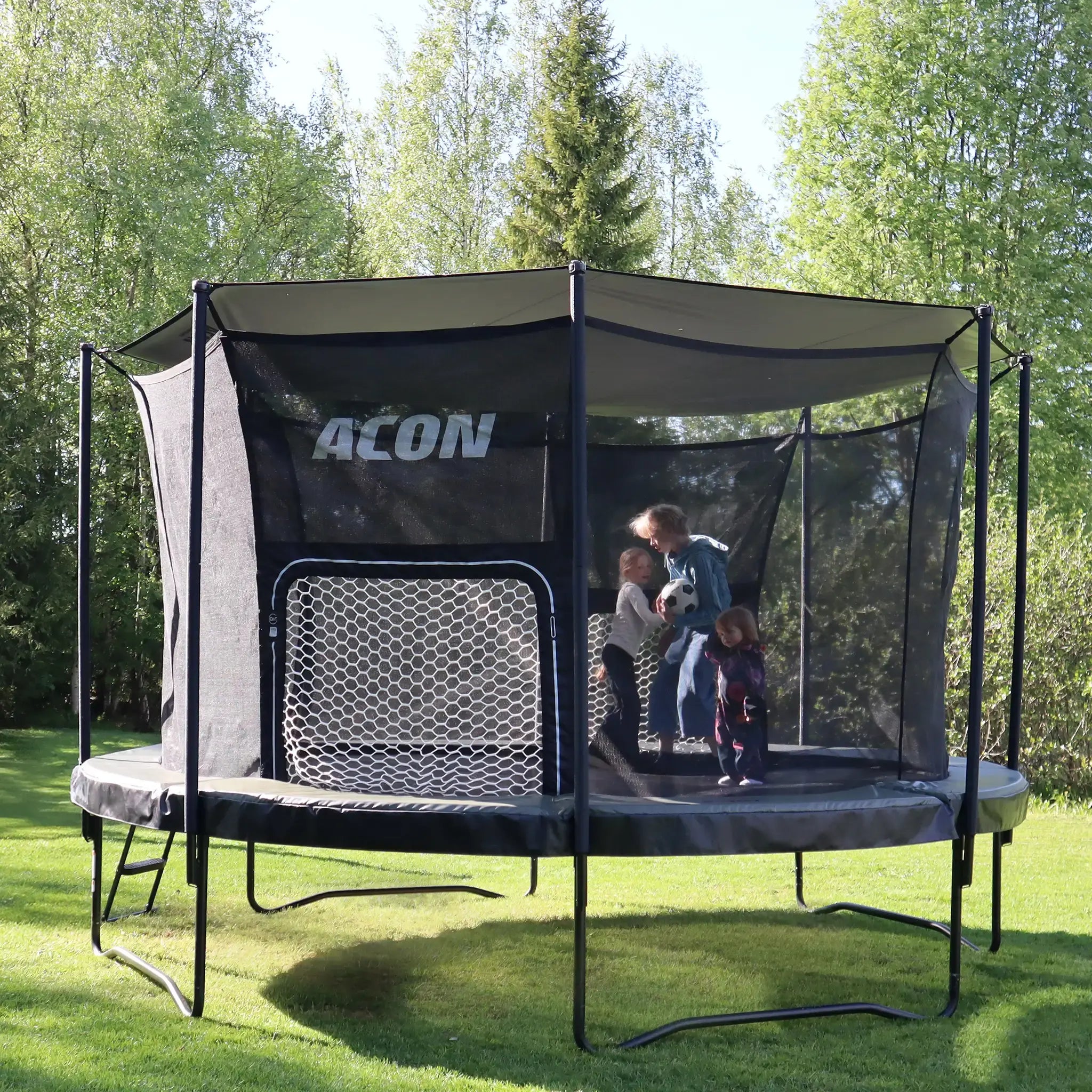 Round Acon air trampoline with sun shade and soccer goal on a grass with three kids playing on it. 