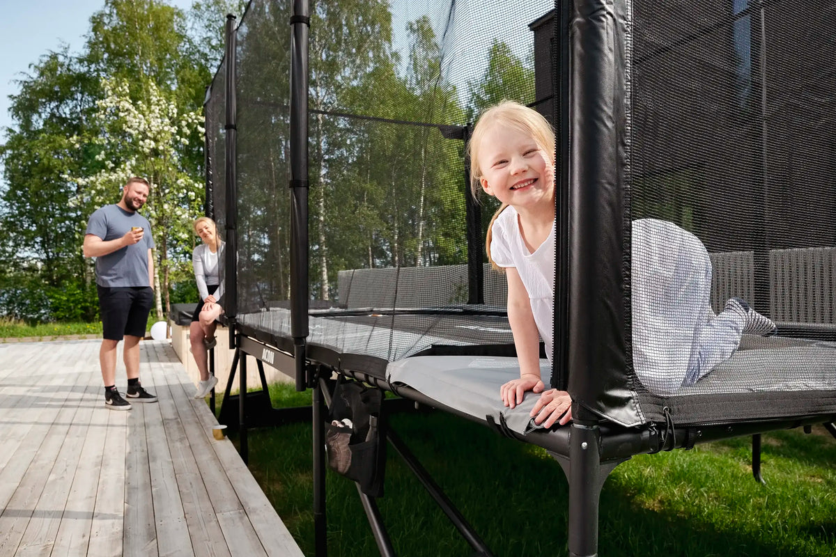 A little girl peeks through the Acon X trampoline netting, as her parents stand watching behind.