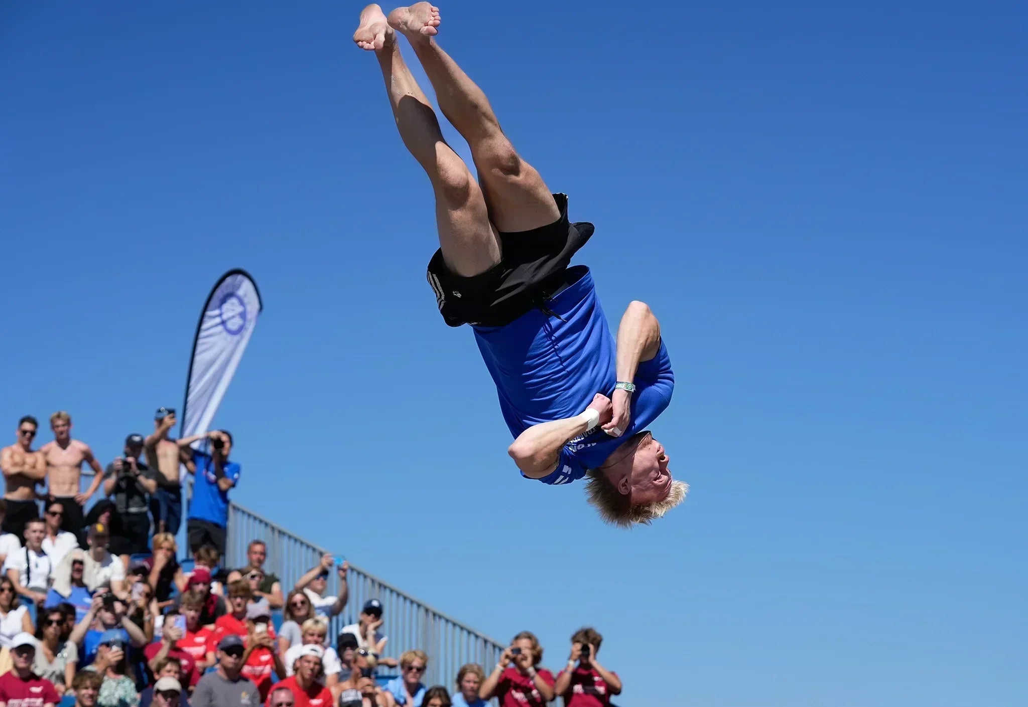 Male athlete flips in the air on trampoline with blue sky and cheering crowd in the background.