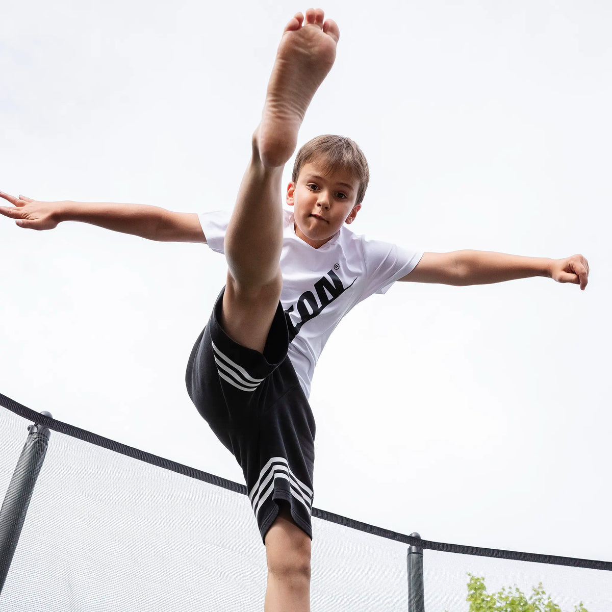 A child wearing a white Acon t-shirt performing a high kick on a trampoline, with one leg extended above his head.