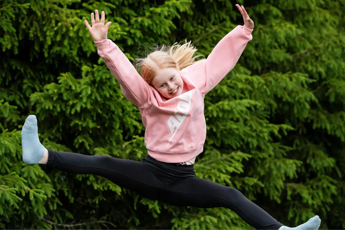 A young girl in a pink Acon sweatshirt jumps on a trampoline with arms up and legs in a split.