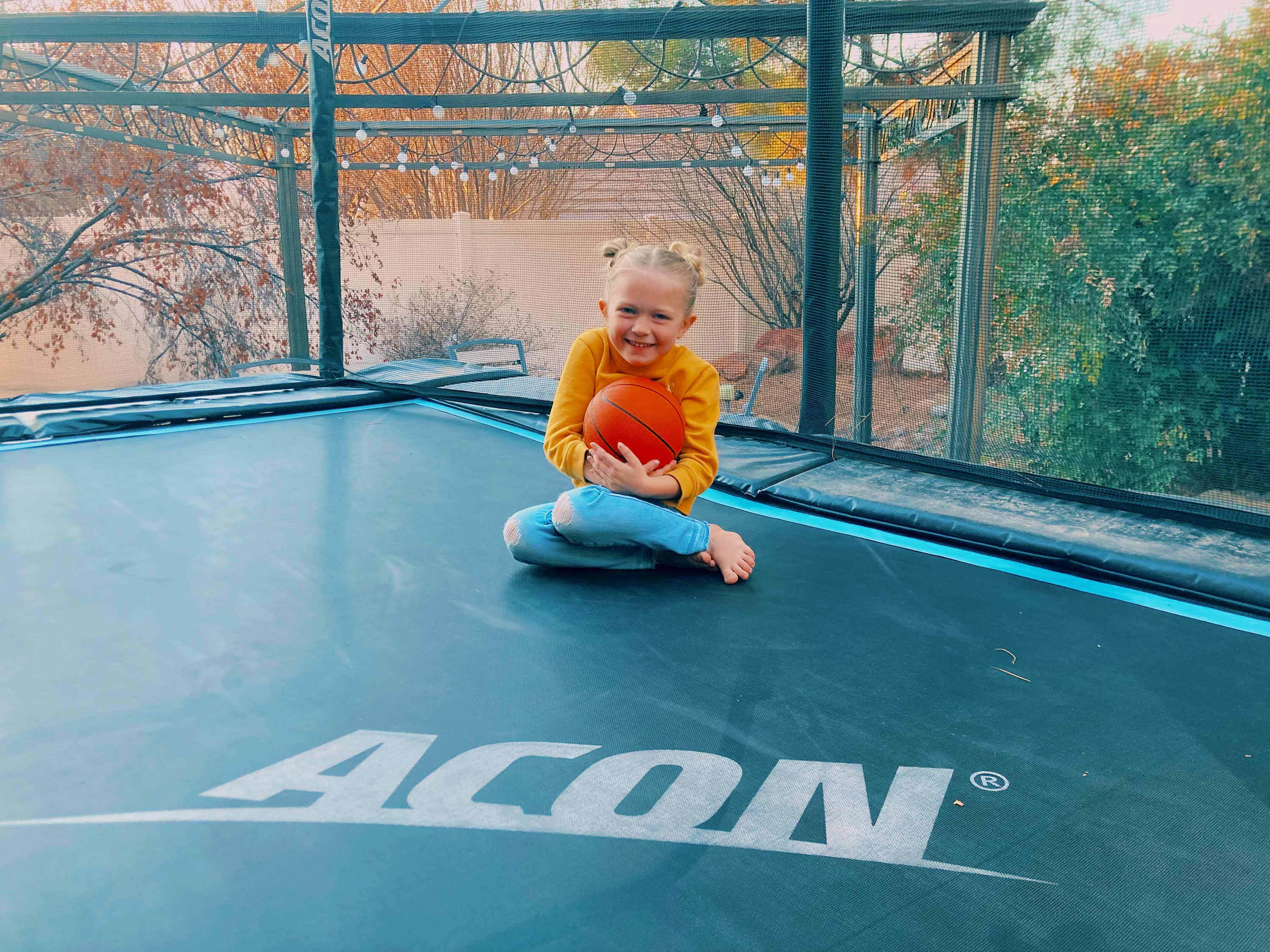 A happy girl sitting on a trampoline and holding a basketball in her arms