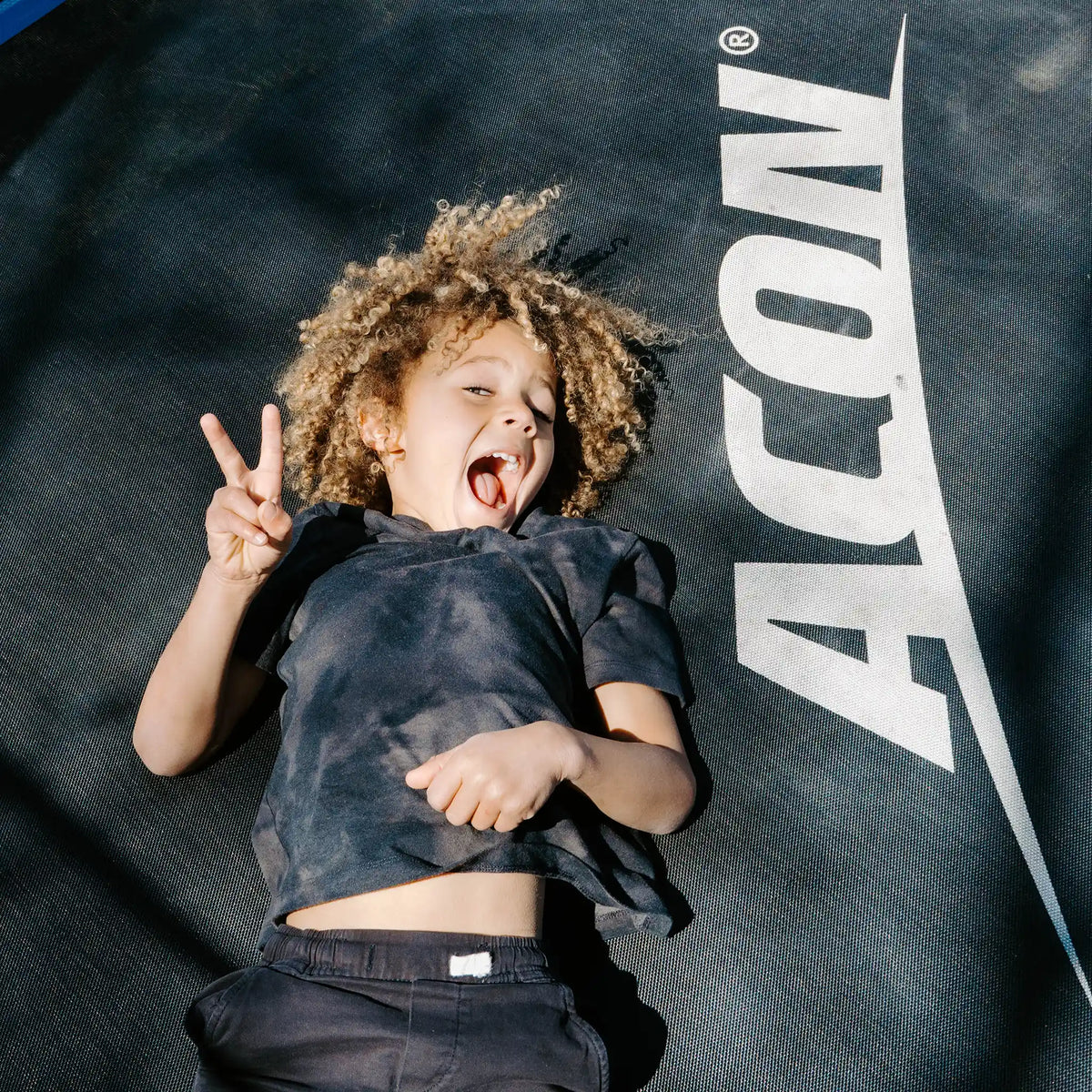 Young child smiling and laying on Acon trampoline and showing peace sign.