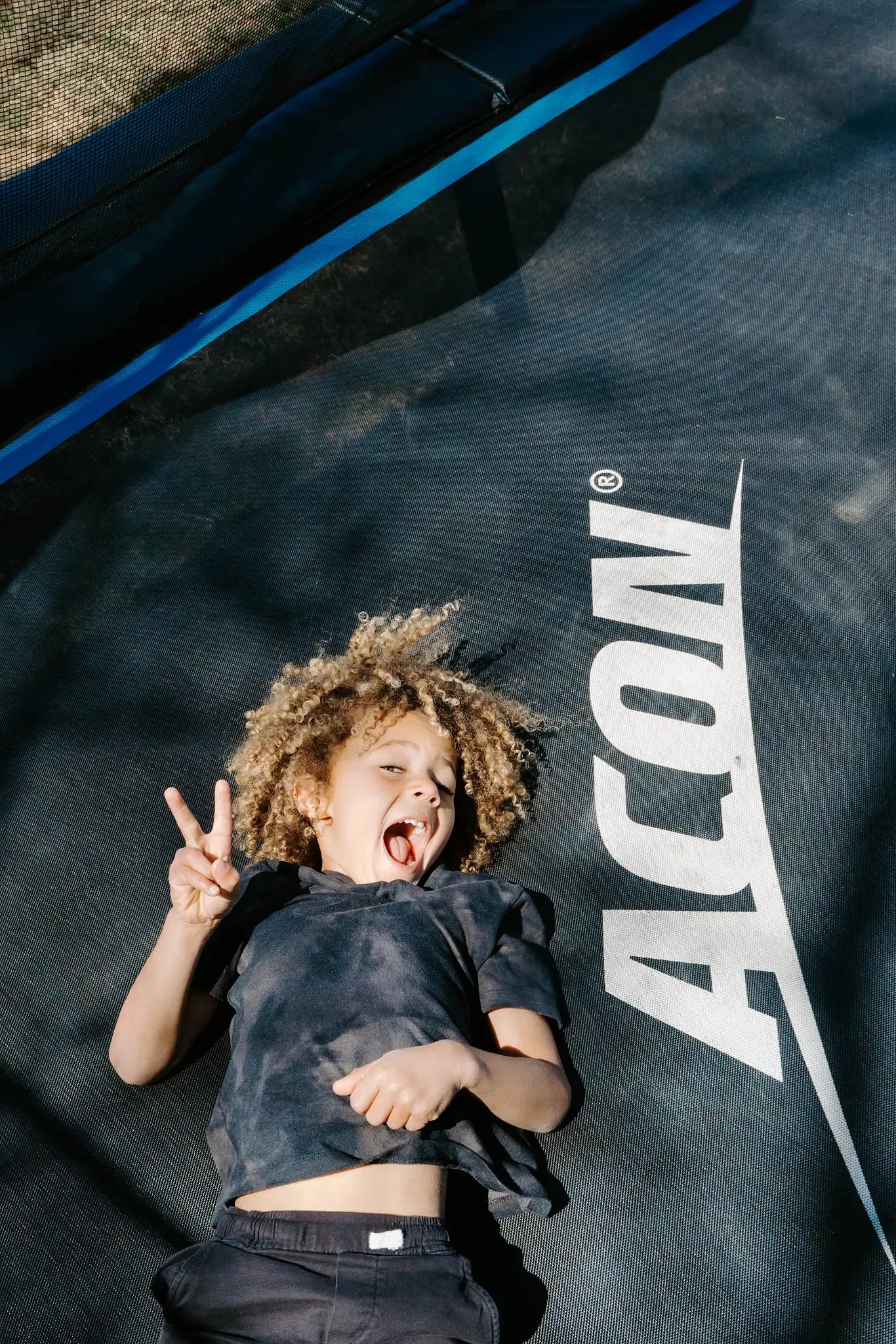 Young child smiling and laying on Acon trampoline and showing peace sign. 