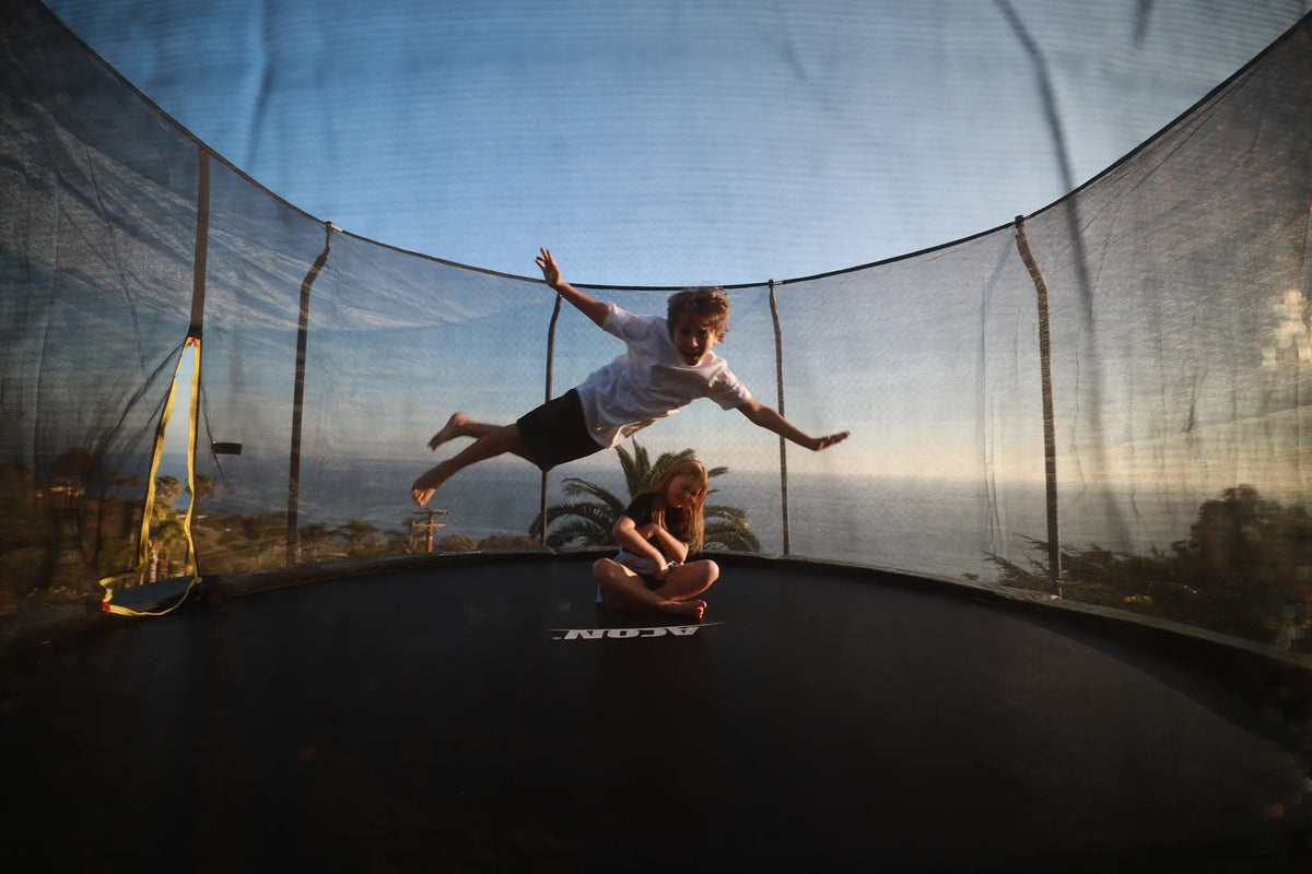 Two children on an ACON trampoline, one jumping while the other sits on the mat.