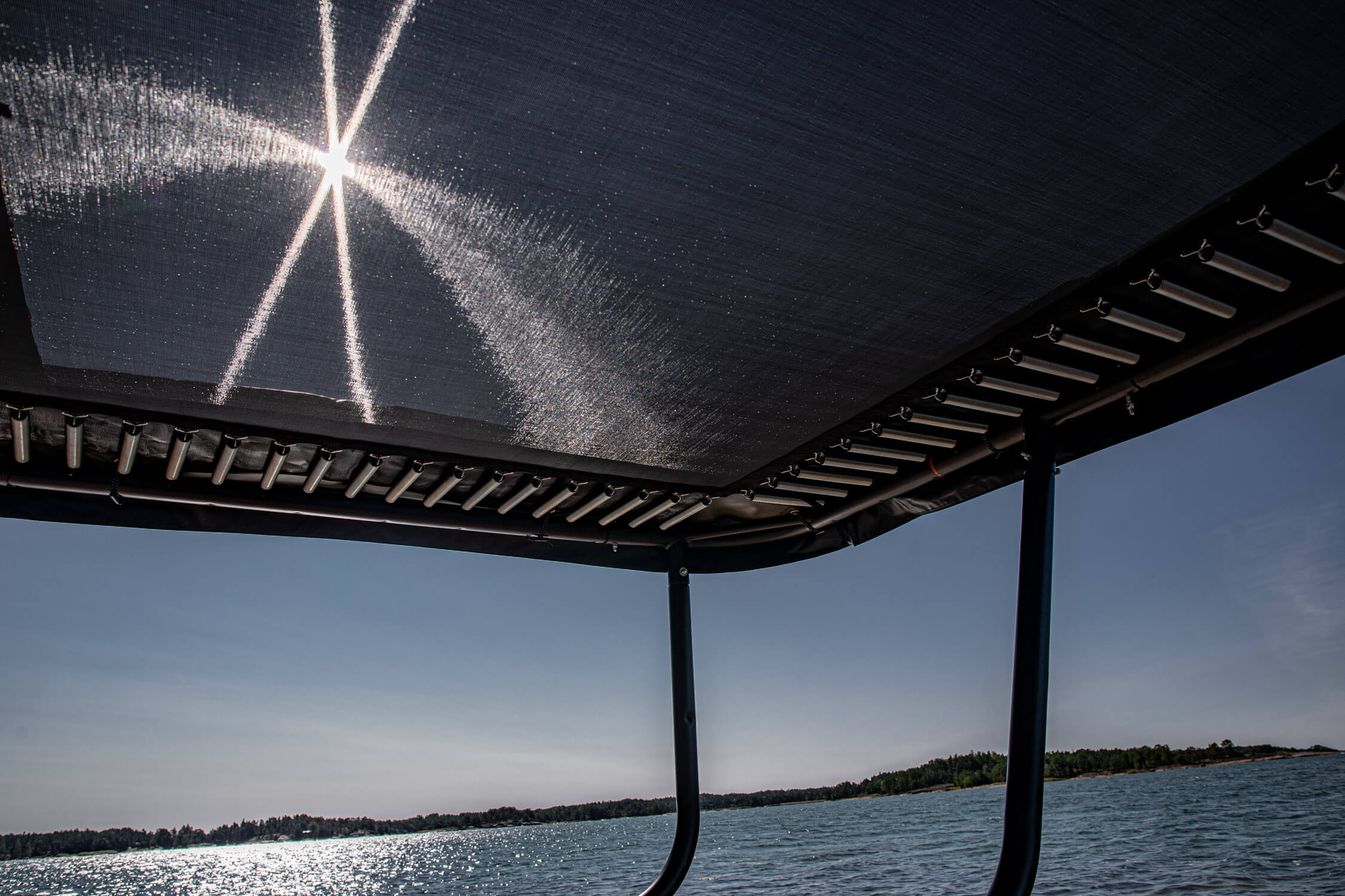 A trampoline pictured from worm's eye view. Sun rays are coming through the trampoline mat and there's a lake in the background