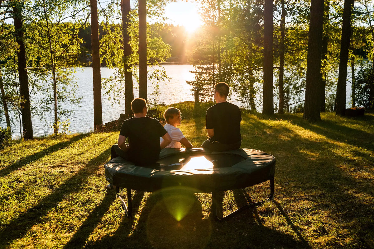 Three boys sitting on an Acon trampoline by a lakeside at sunset, surrounded by trees.