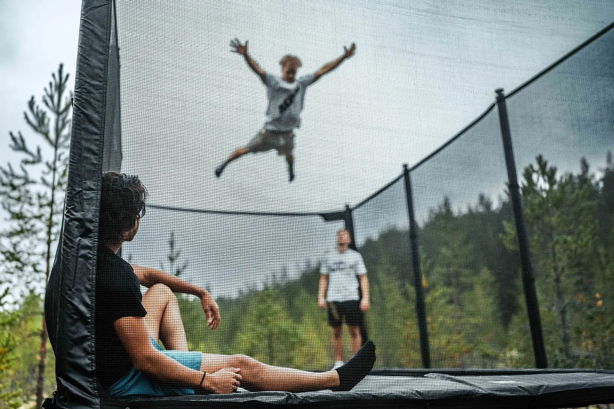 Three people using a Acon trampoline outdoors, with one person jumping high in the air and others watching inside the safety enclosure.