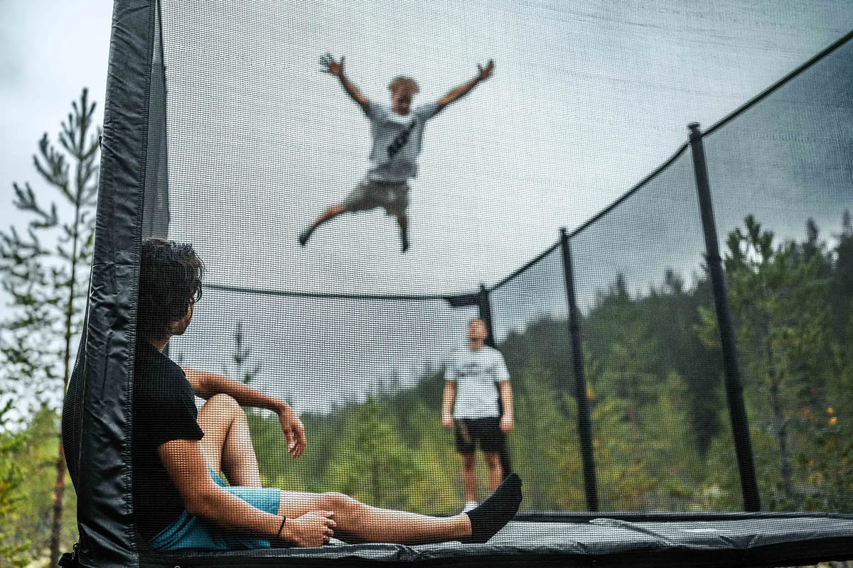Three people on an Acon trampoline outdoors, with one jumping high in the air and others watching.