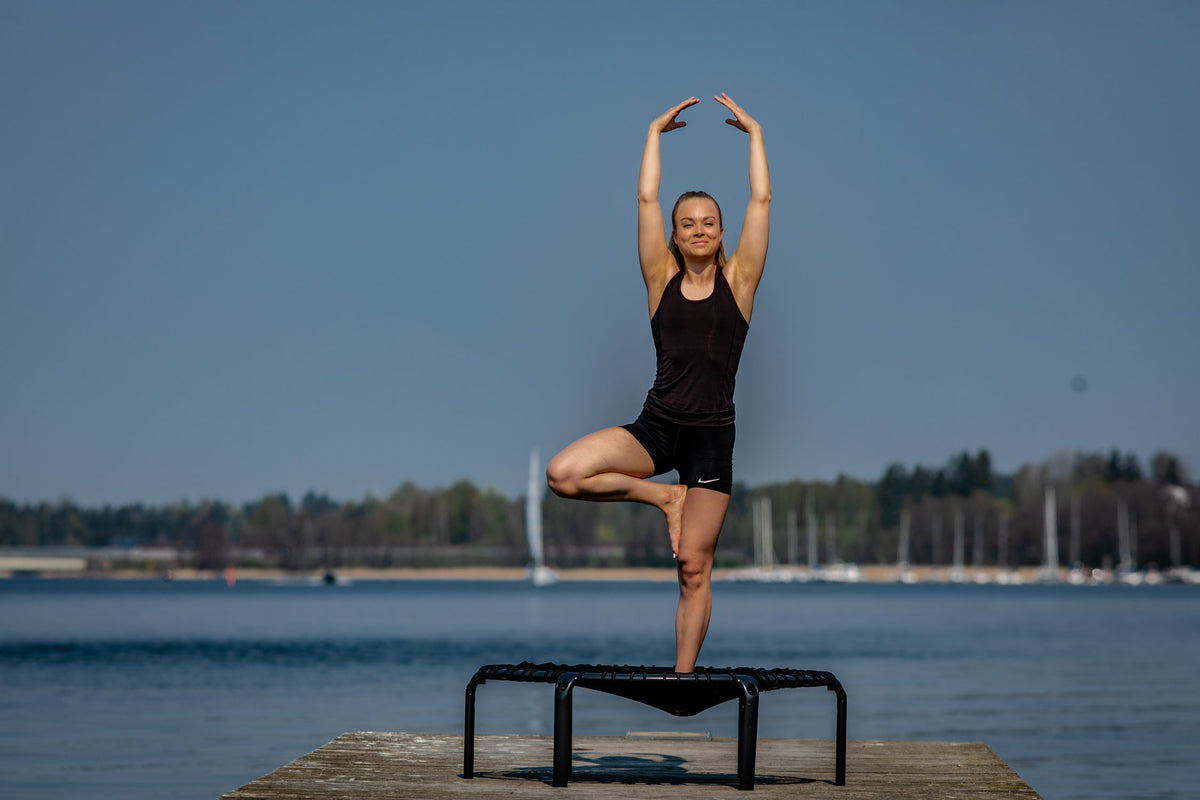 A woman practicing tree pose and doing trampoline yoga on an ACON rebounder trampoline outdoors