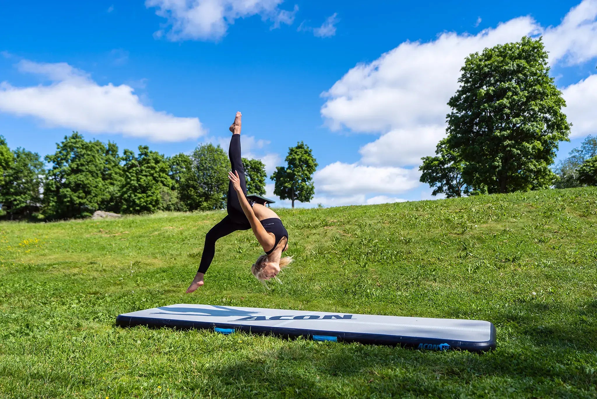 Gymnast practicing an aerial skill on an ACON Airtrack in a grassy outdoor training area.