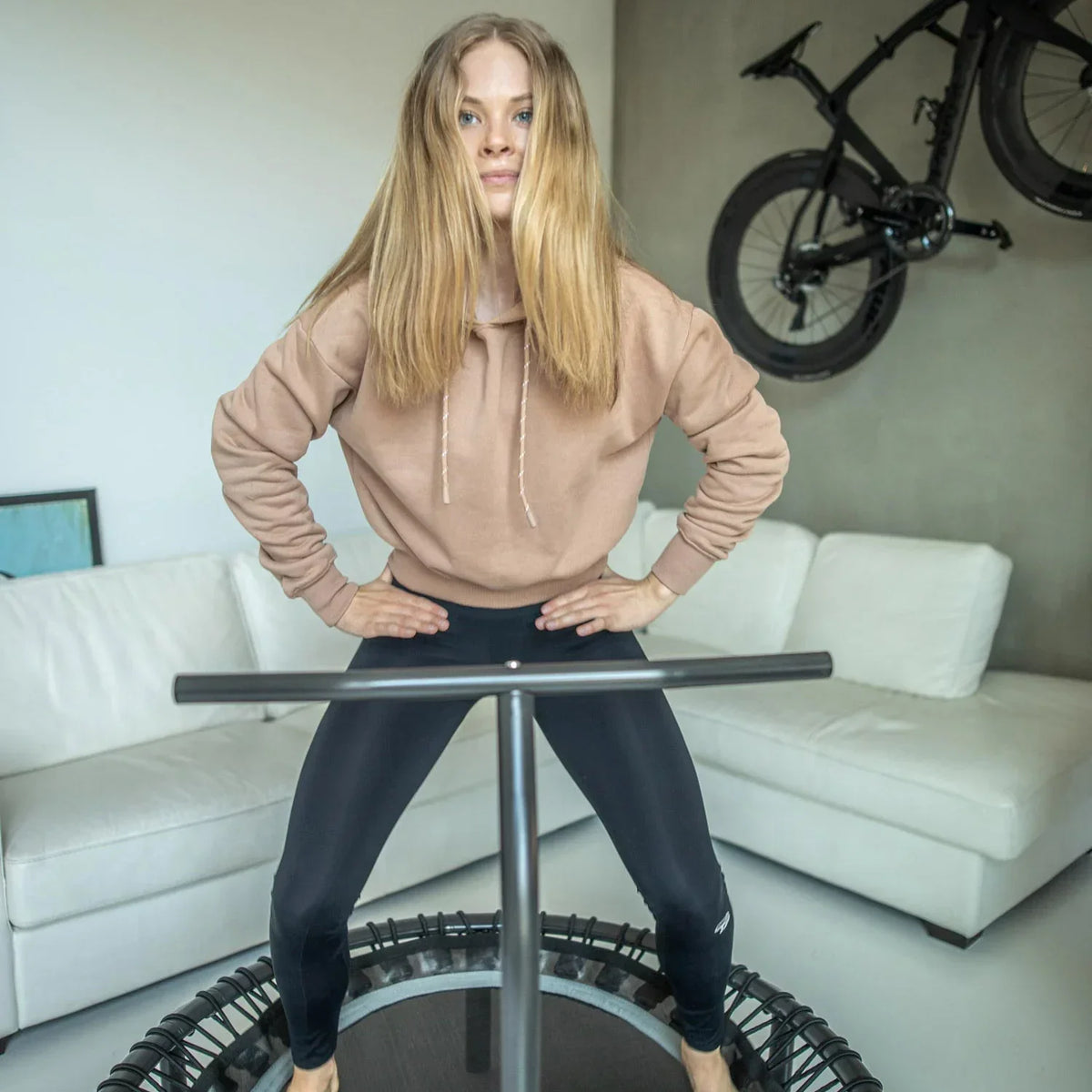 Woman standing on a rebounder with a handlbar, infront of a white couch and a bike on the wall.