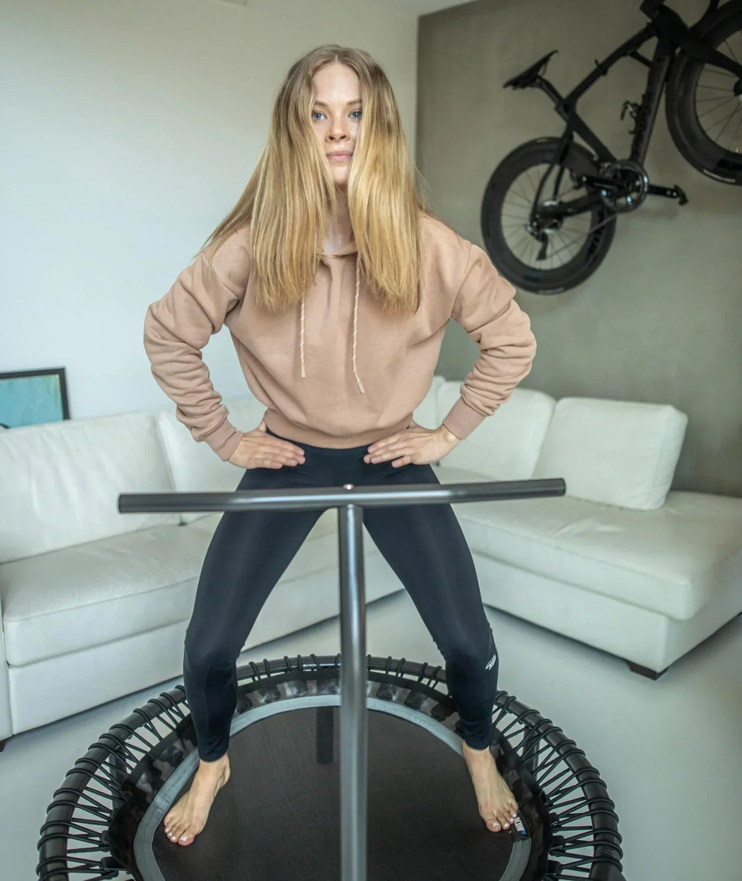Woman standing on a rebounder with a handlbar, infront of a white couch and a bike on the wall.