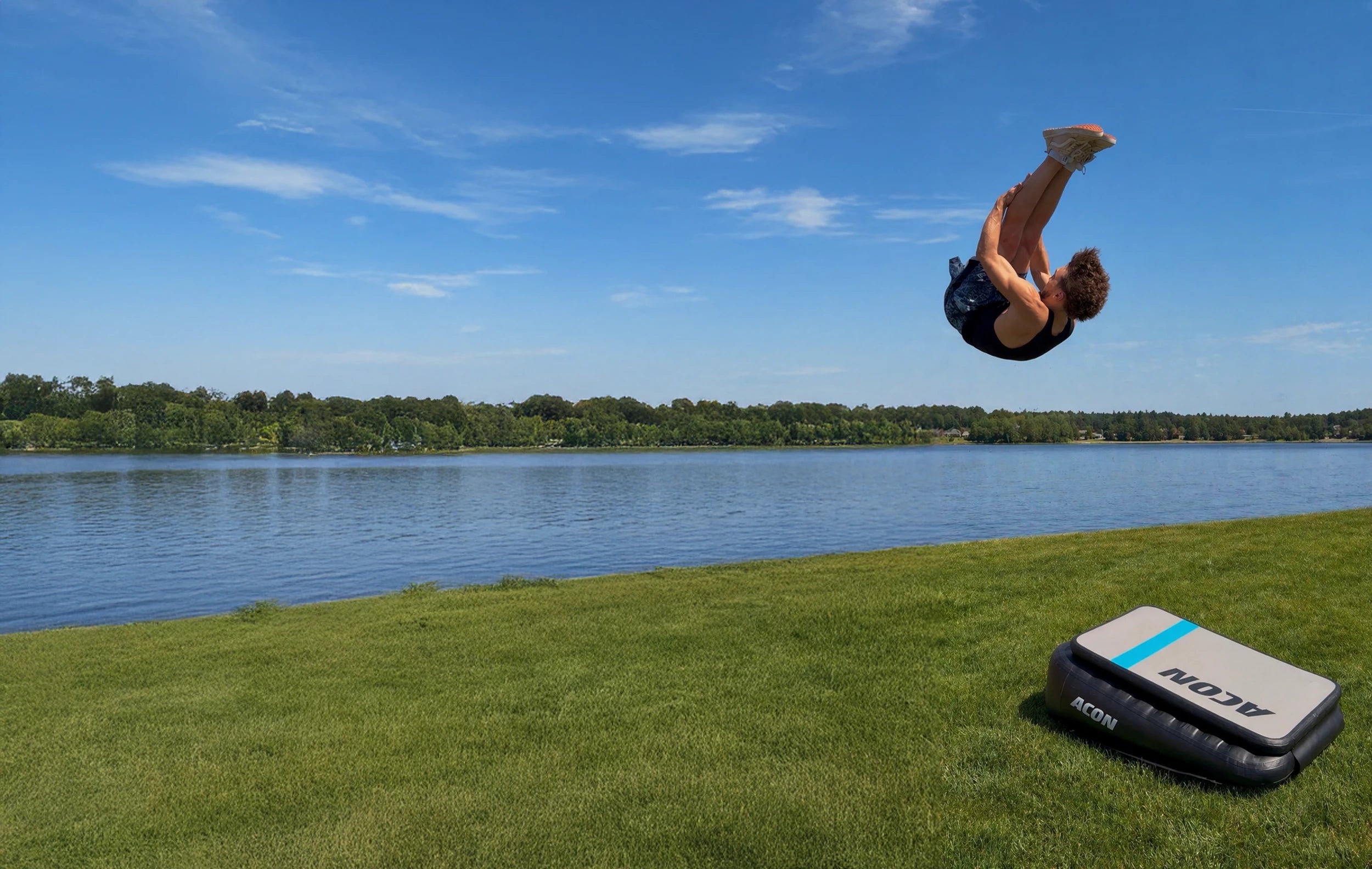 Man jumping a Pike Front on an Acon Airboard by a riverside, the Airboard set on grass.
