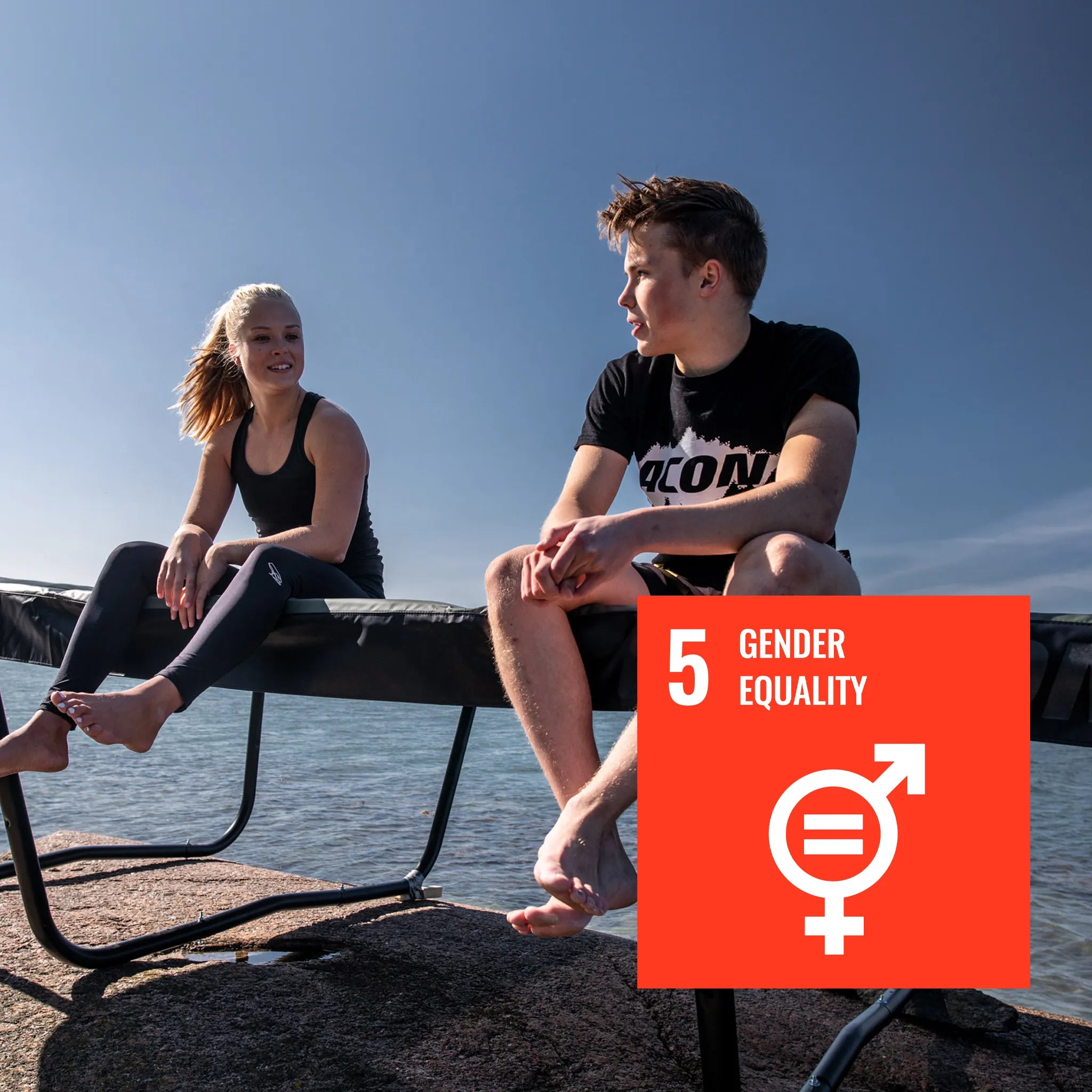 A woman and a man are sitting on an Acon HD trampoline. The image features the UN sustainability mark 5 - Gender Equality.