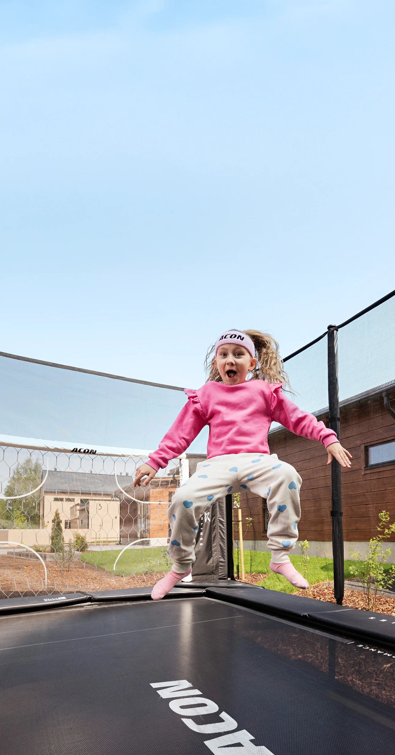 A joyful young girl in a pink shirt and Acon headband jumps on an Acon X Trampoline, her happy expression showing pure fun. Houses and blue sky behind her.