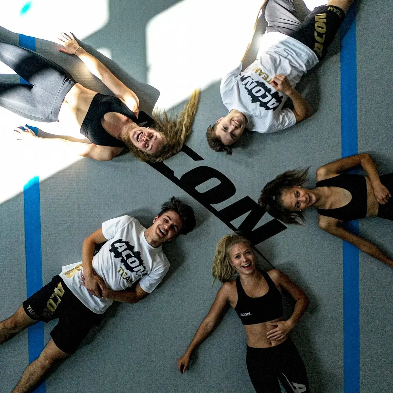  Five people in athletic wear lie on a big ACON airtrack, around a large “ACON” logo, smiling up at the camera.
