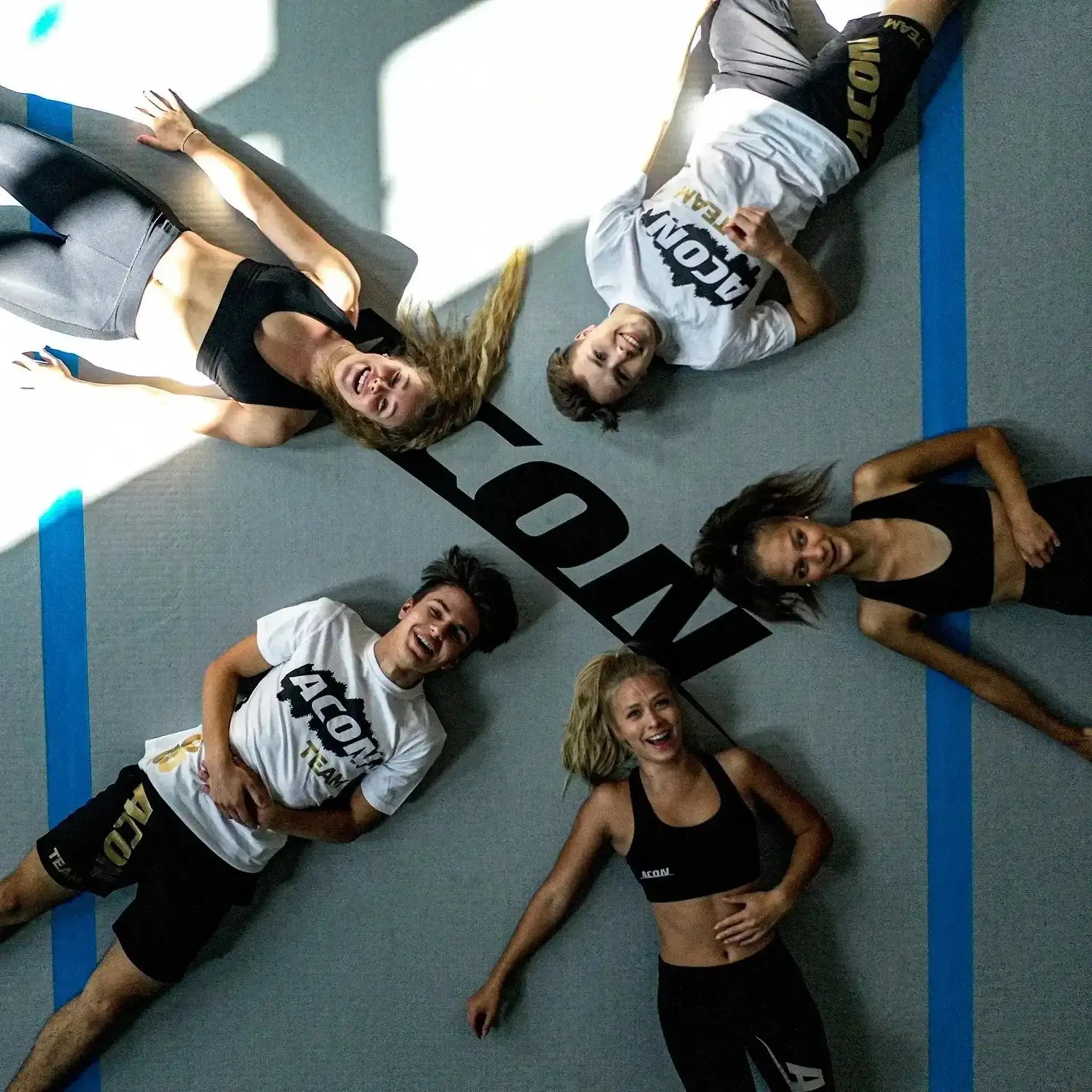  Five people in athletic wear lie on a big ACON airtrack, around a large “ACON” logo, smiling up at the camera.