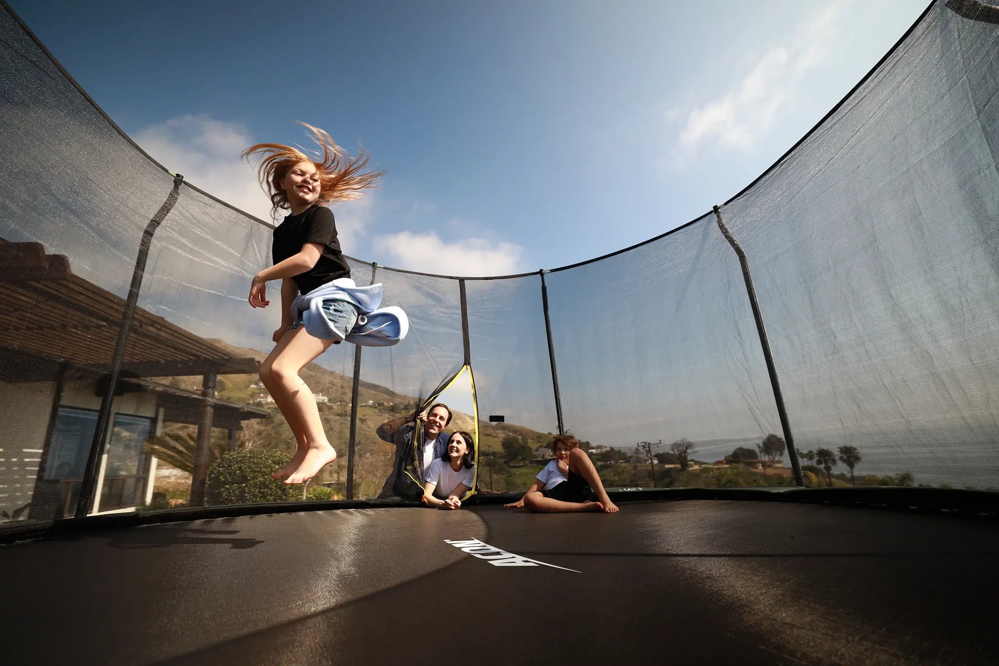 Girl jumping on an Acon trampoline while family watches inside the enclosure.