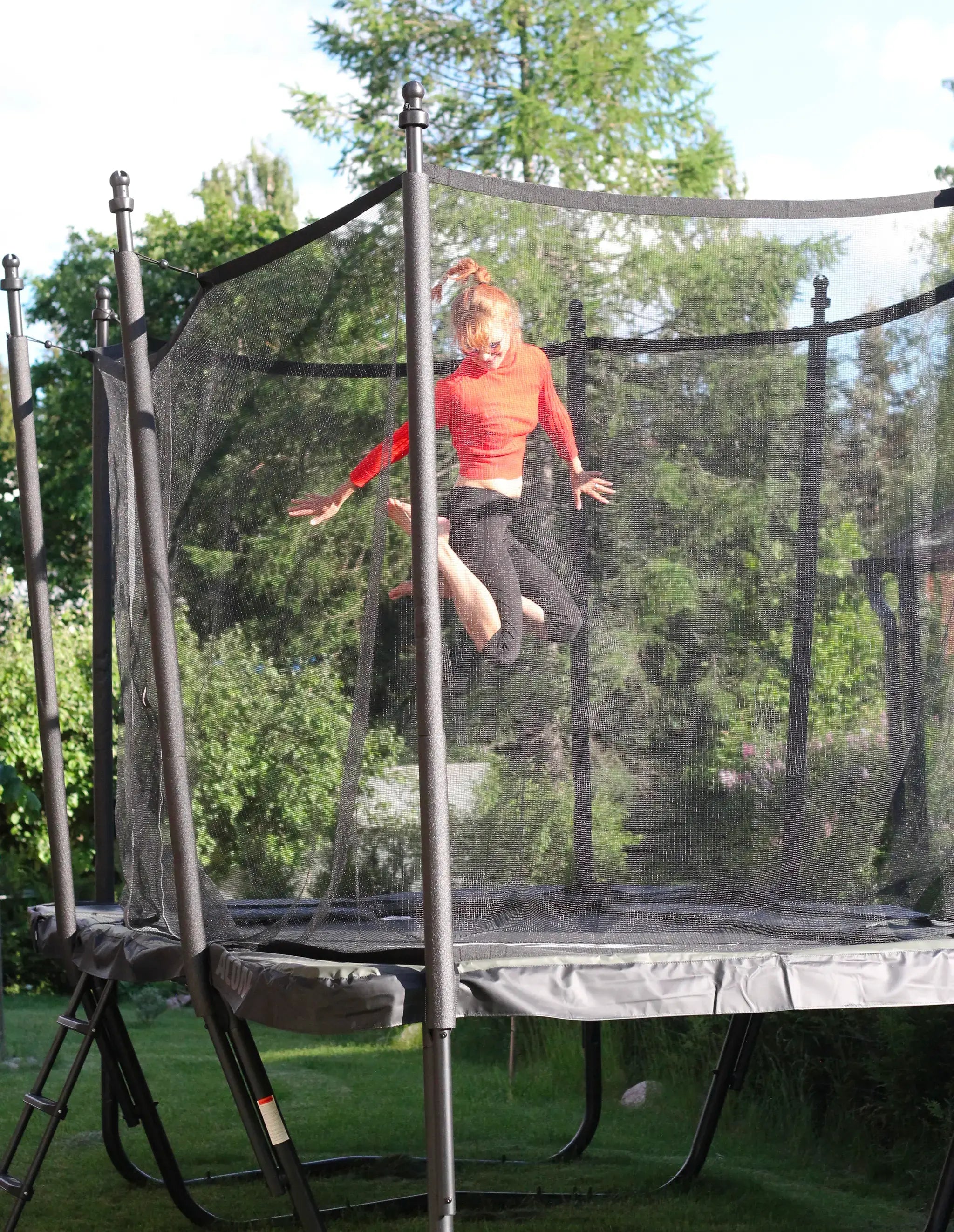 A teenager in a red shirt mid-air while jumping on a trampoline in a green backyard.