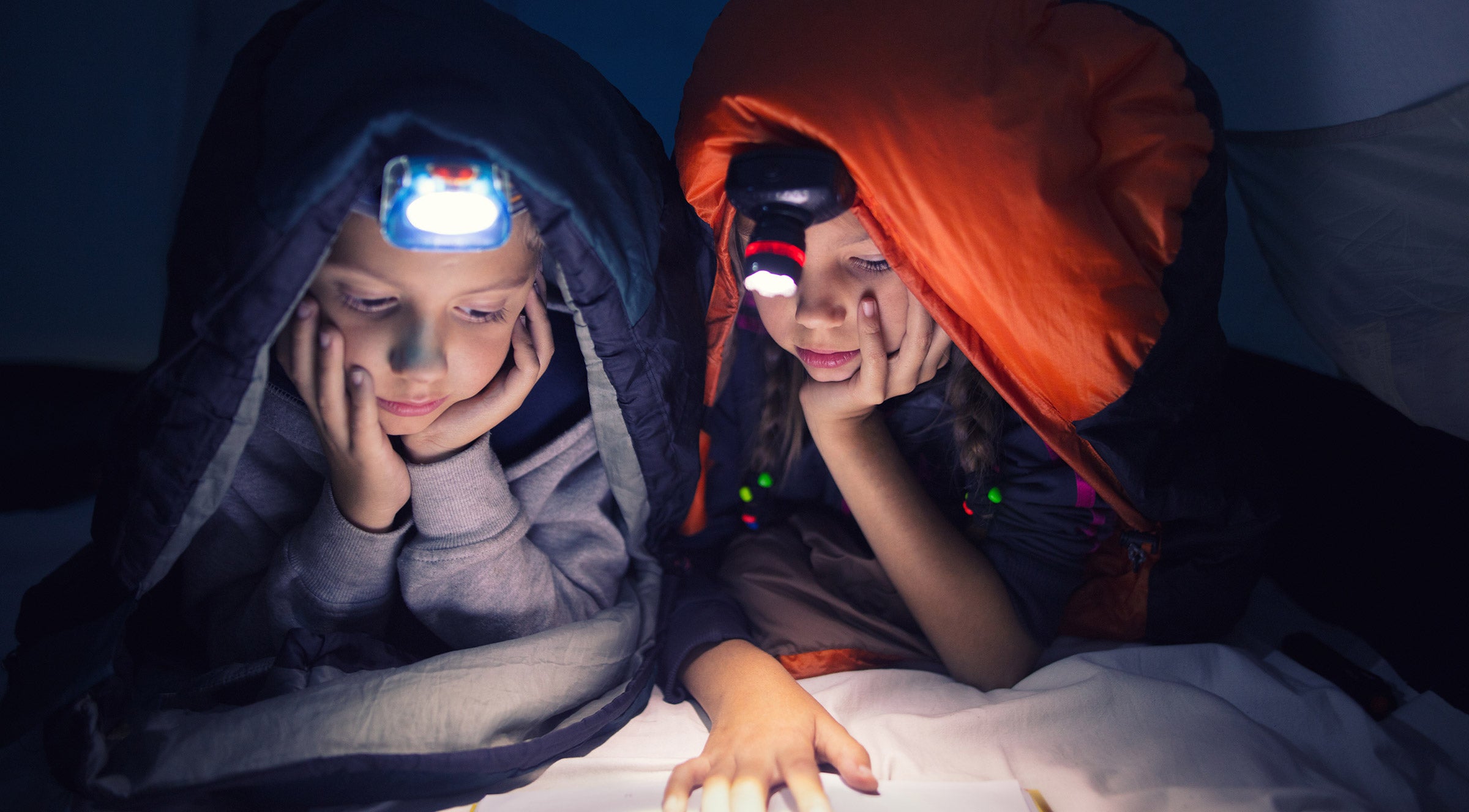 Two girls in a tent reading by headlamp light.