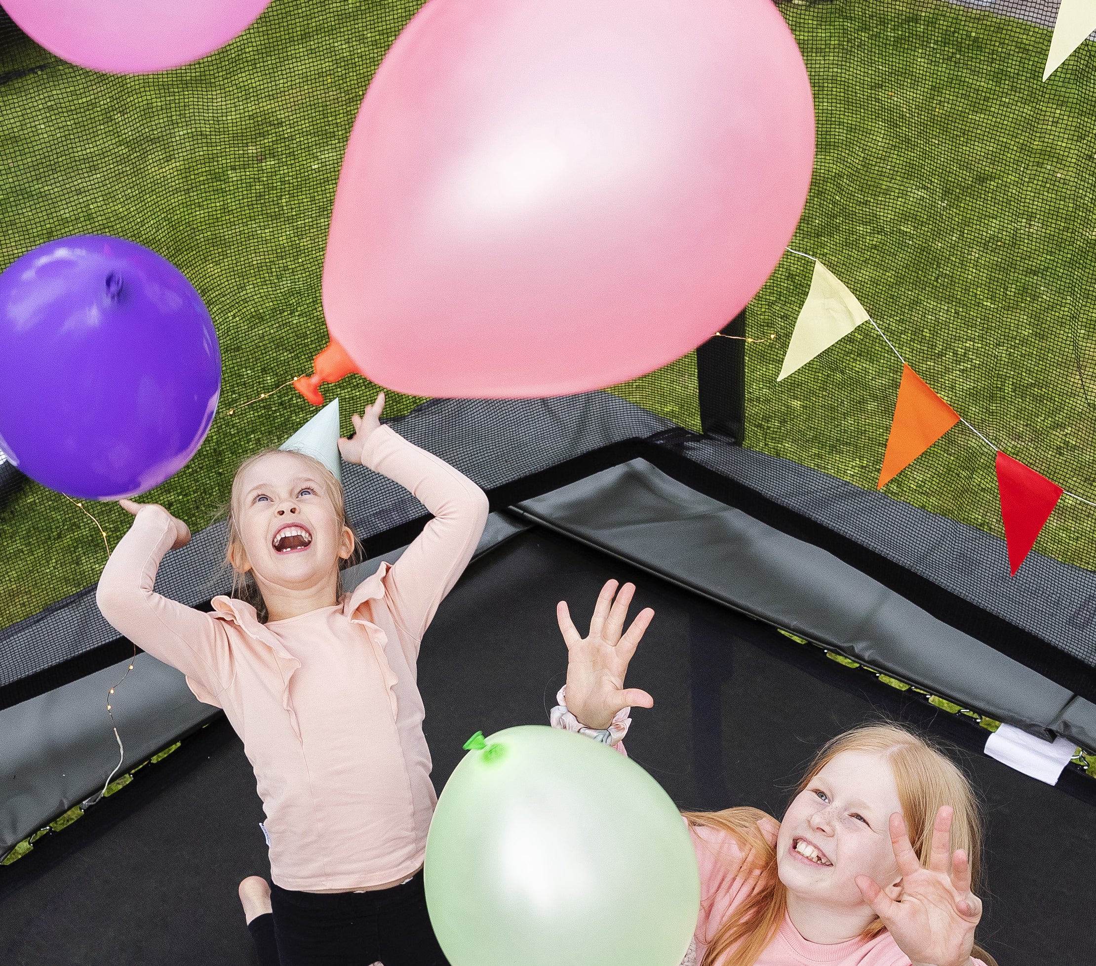 Two girls on a rectangular trampoline playing with birthday balloons.