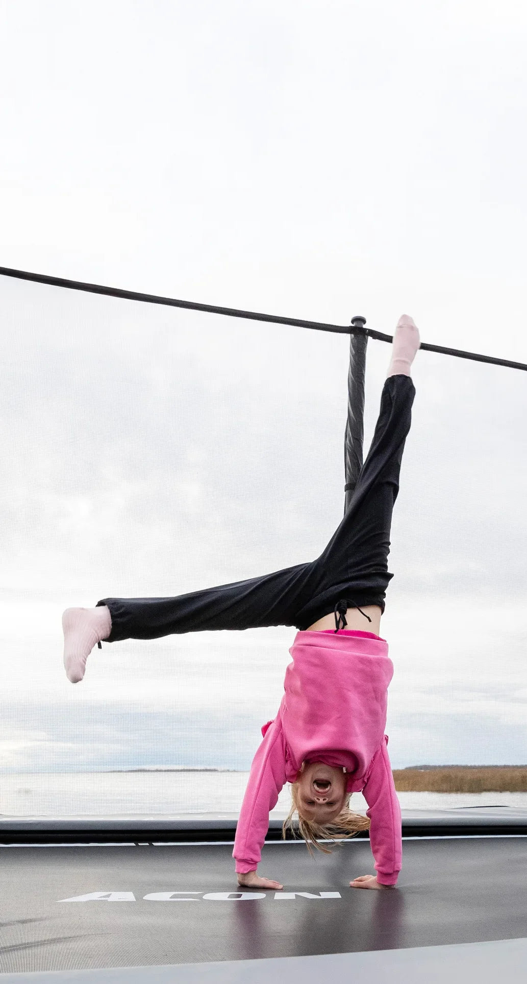 A joyful girl wearing a pink shirt is doing a handstand on an Acon X trampoline.