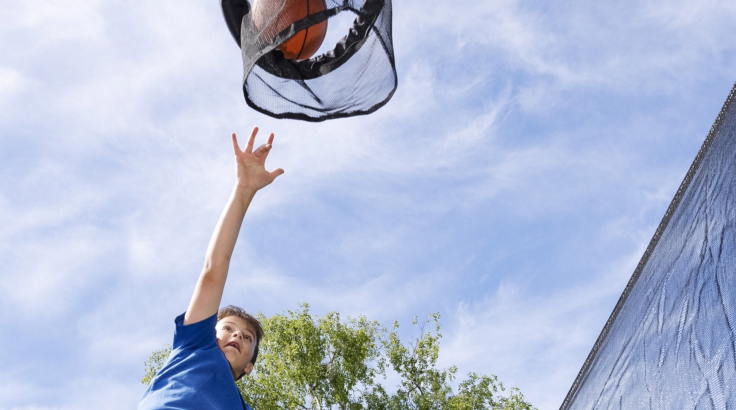 A boy on a trampoline jumping and throwing a basketball into the hoop.