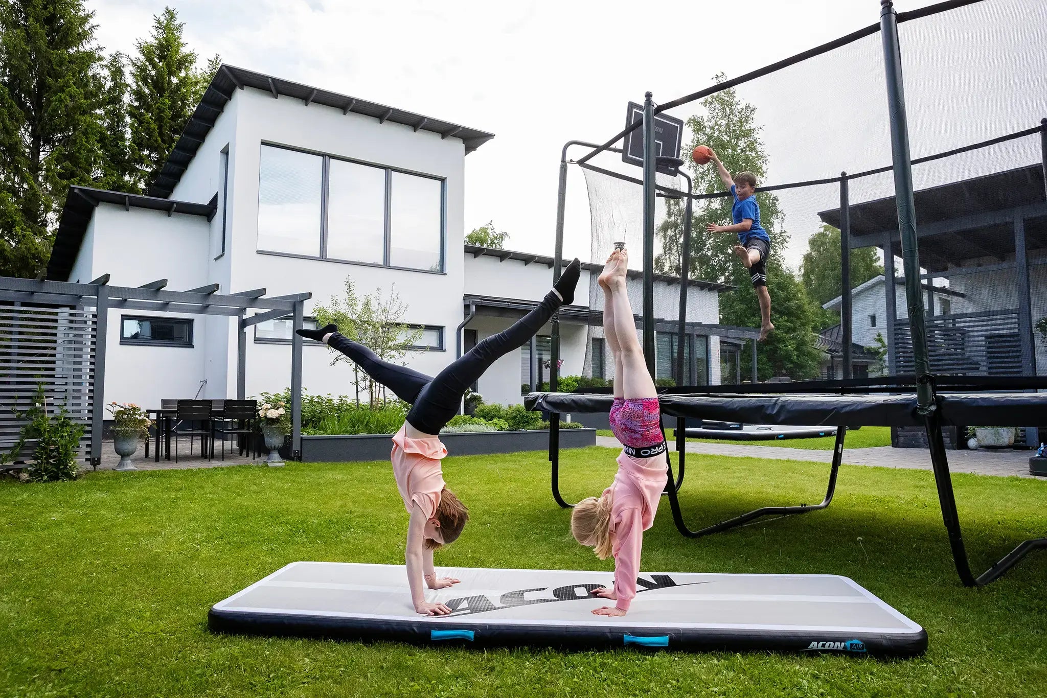 Three kids playing around with rectangular Acon trampoline and a air track, two girls are doing tricks on the ground while the boy is jumping and playing basket ball. 