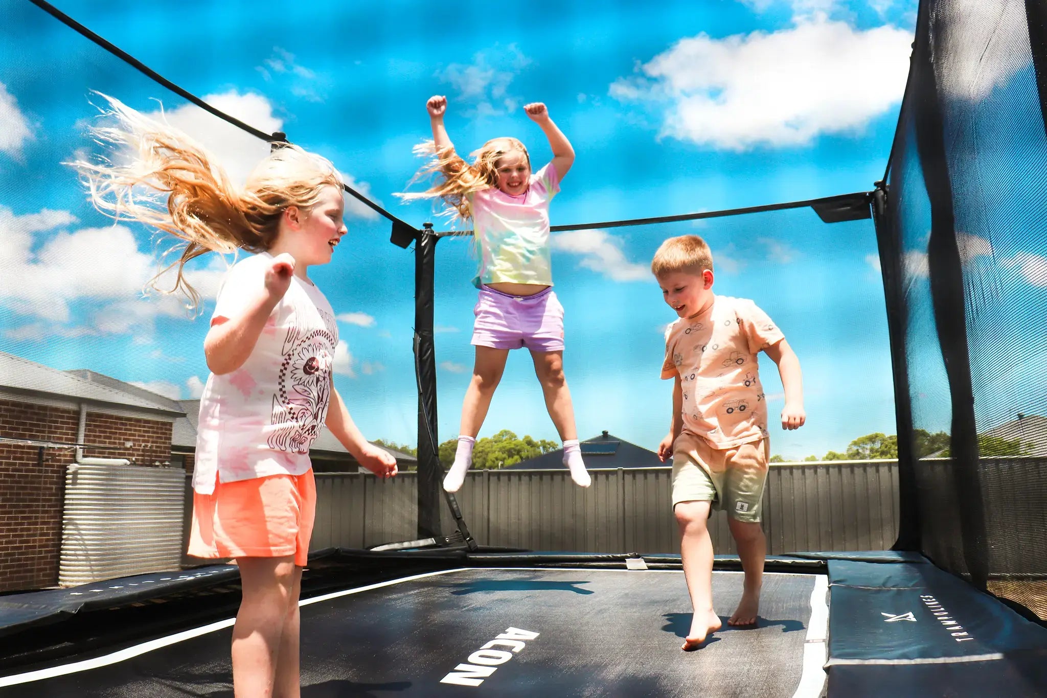Two young girls and a boy jumping happily on a rectangular Acon trampoline. 