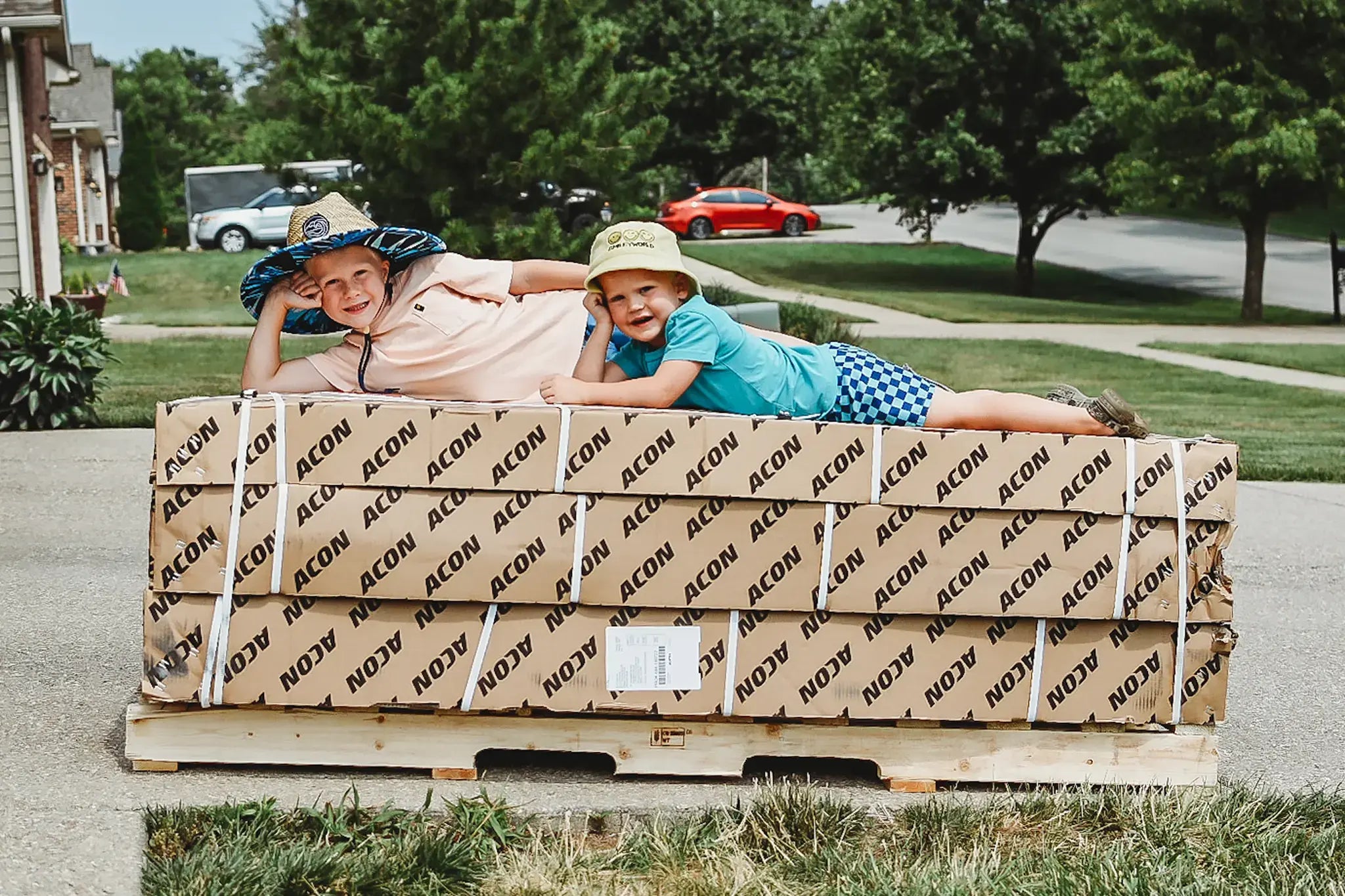 Two kids smiling and lying on top of a boxed Acon trampoline delivery on a driveway.