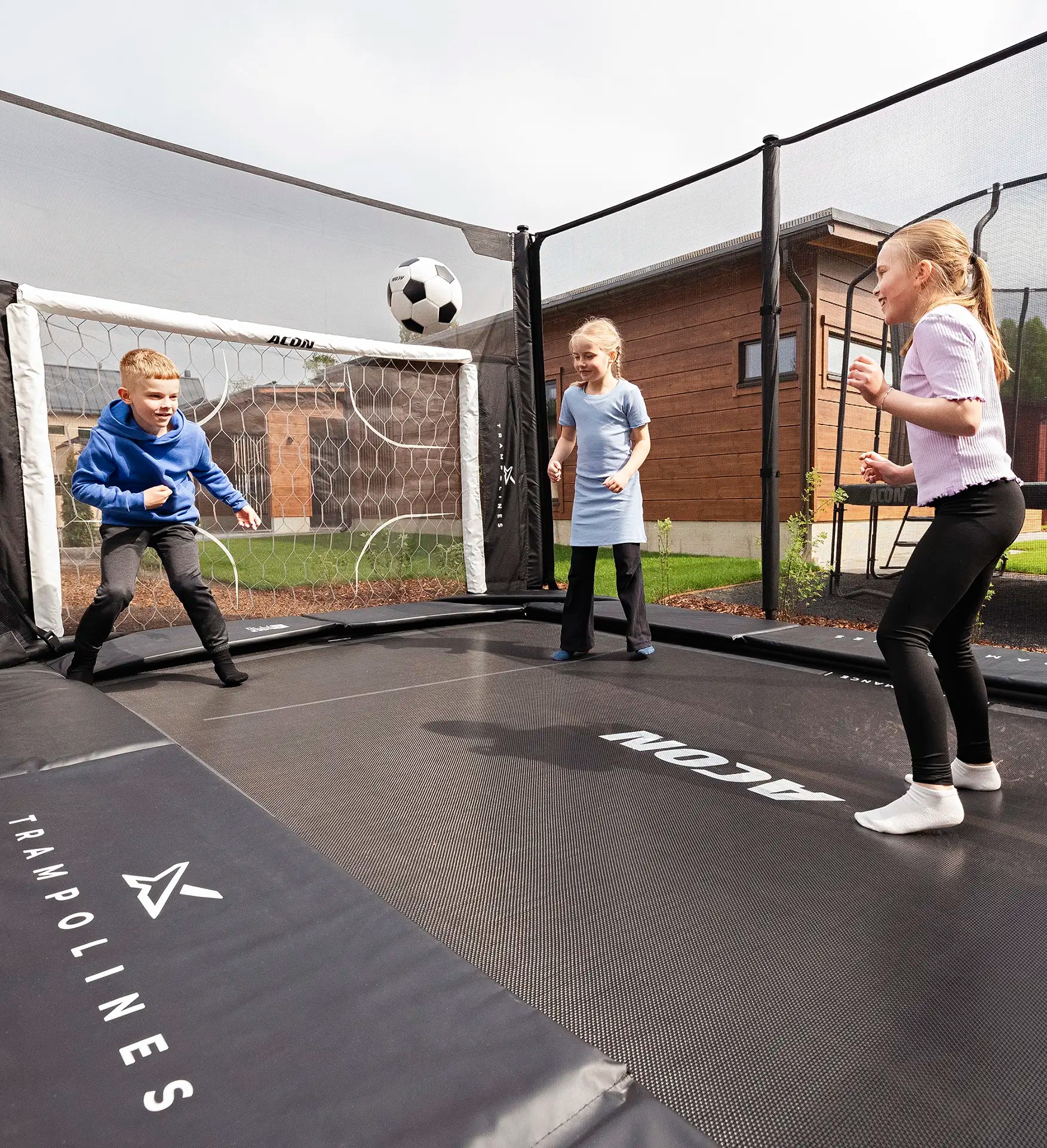 Children playing with a soccer ball on an Acon X 17ft trampoline with a soccer goal in the background.