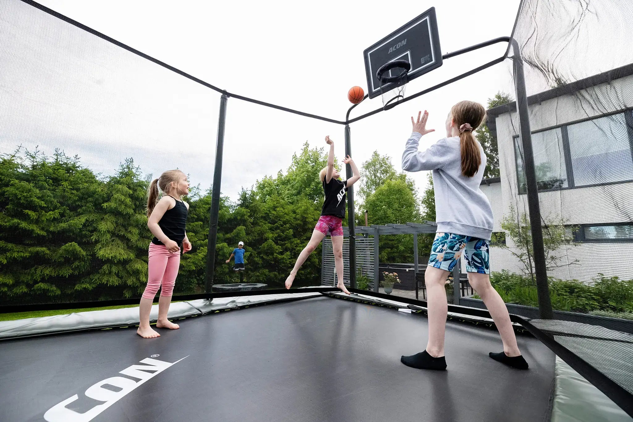 Kids playing basketball on a Acon trampoline with a basketball hoop in a attached to the trampoline's safety net. 
