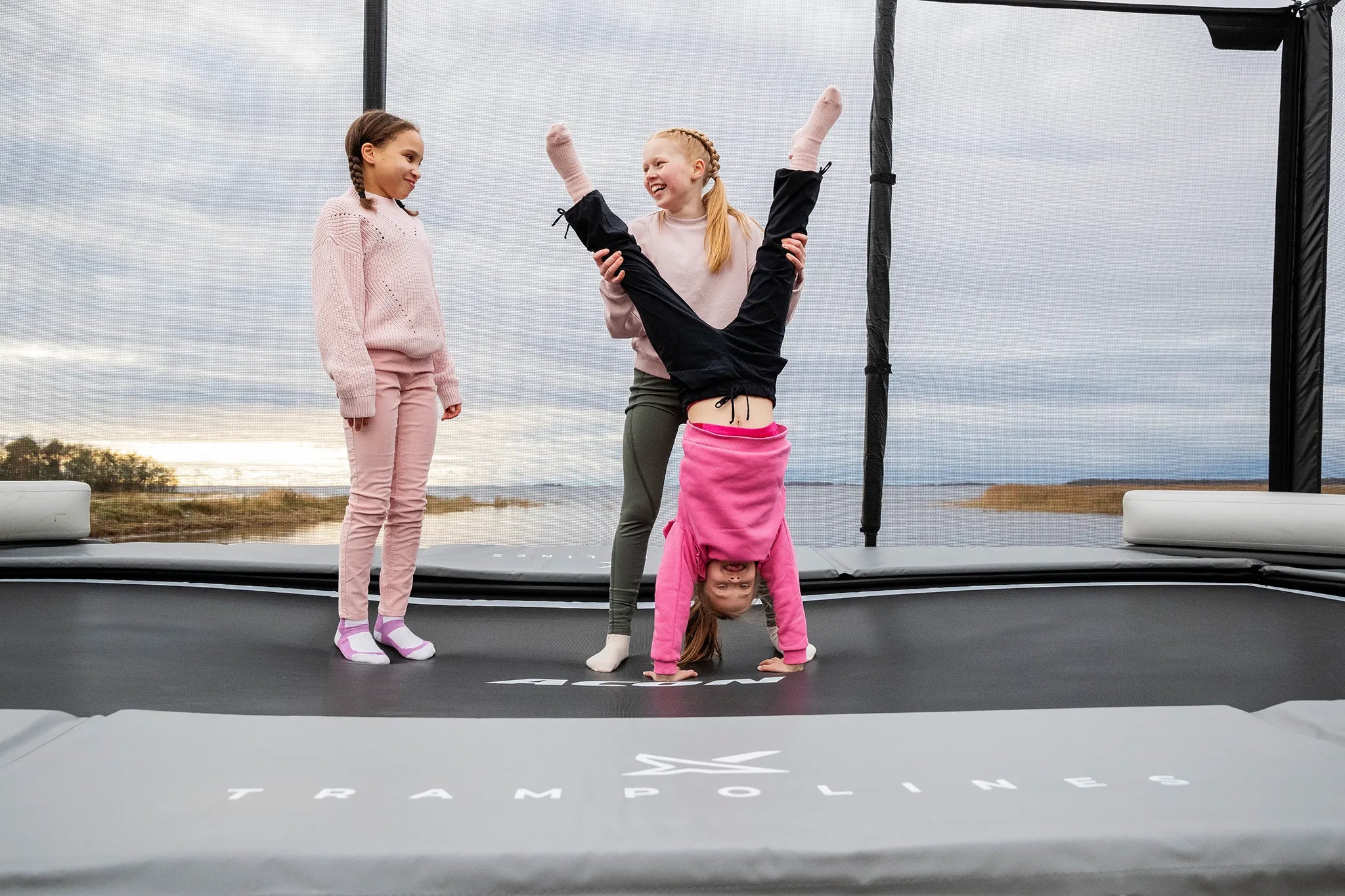 Three girls on a Acon X trampoline, one is doing a hand stand while other one is helping by holding her legs. 
