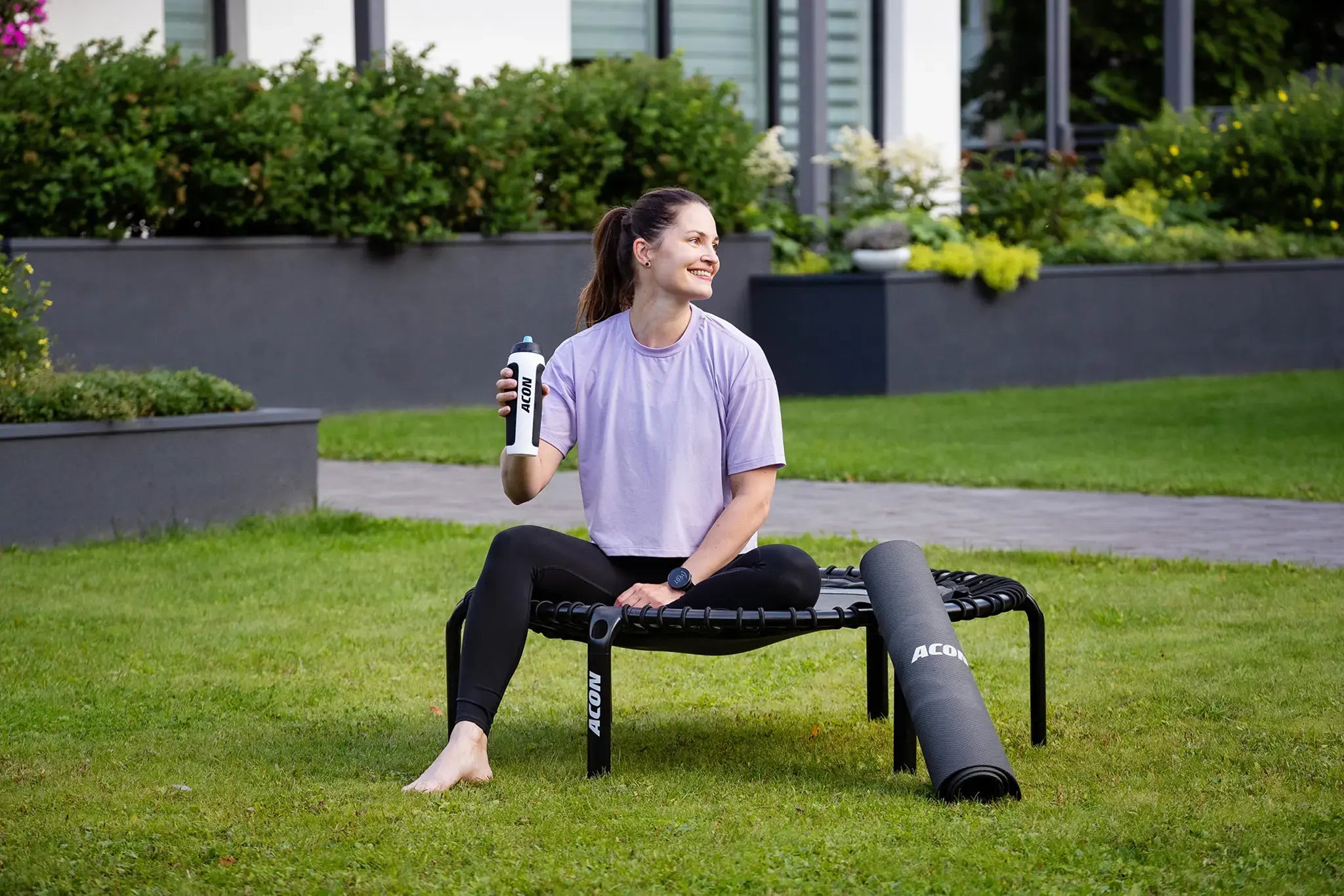 Woman taking a break and smiling while sitting on an Acon rebounder in the garden, holding an Acon water bottle with a fitness mat beside her.