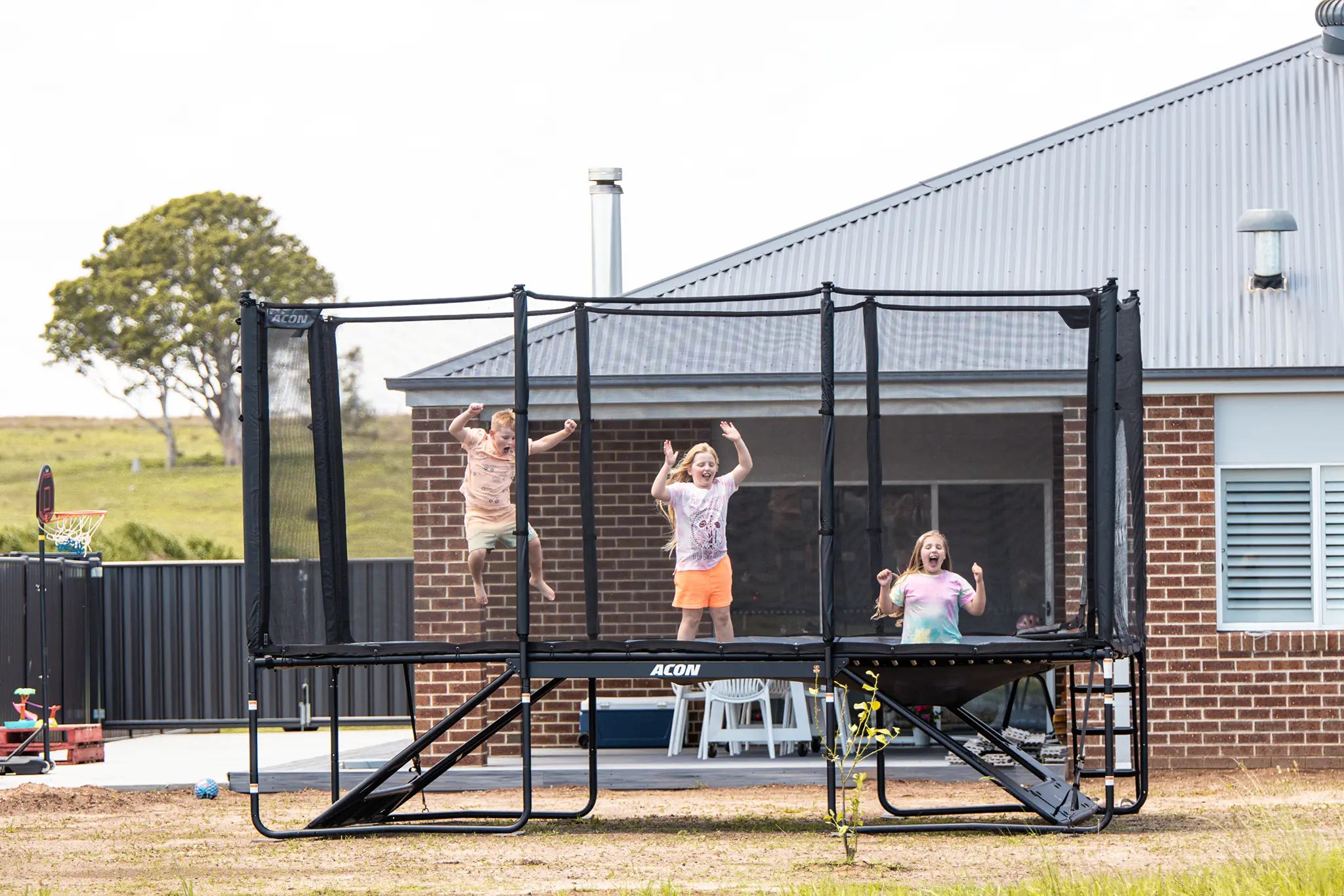 Three children jumping and playing on an Acon X trampoline with a safety net in a backyard next to a brick house.