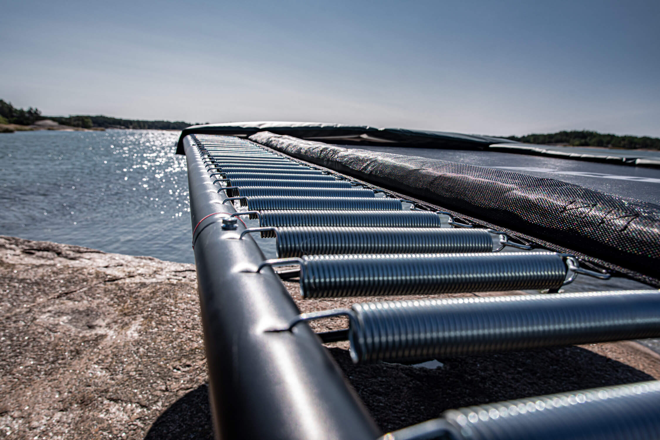 A closeup of ACON trampoline springs. The trampoline is on a beach and there's sea in the background