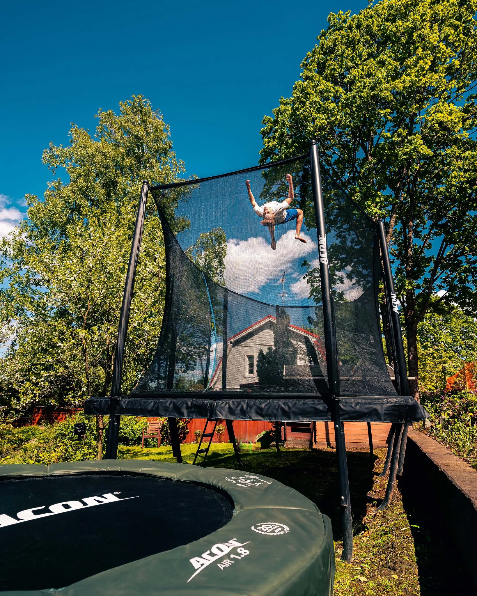 A guy doing a backflip on an ACON rectangular trampoline with net.
