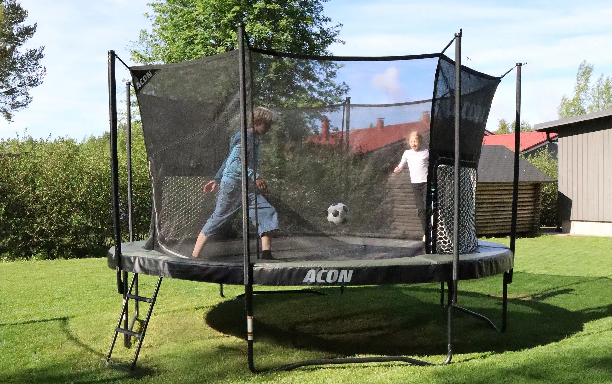 Two kids playing soccer on Acon trampoline with goal net in backyard garden setup.