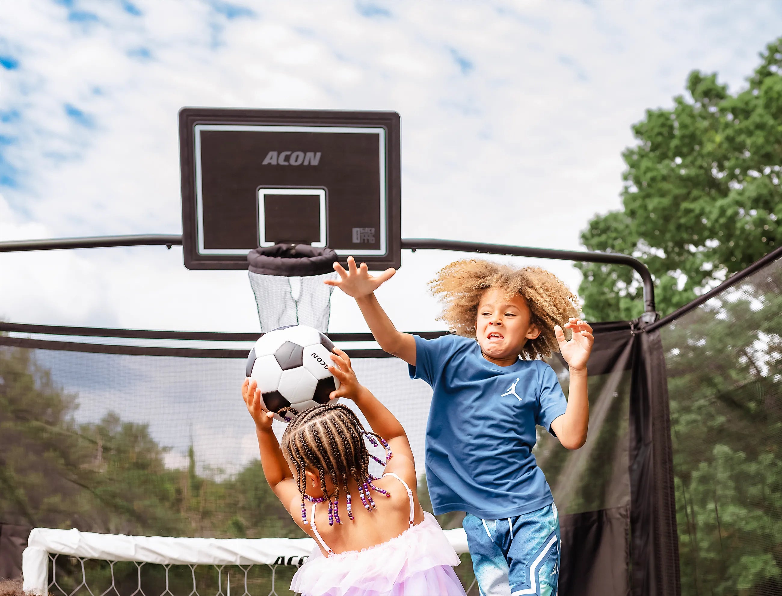 Two children playing with a soccer ball on an Acon X Trampoline with a soccer goal and a basketball hoop.