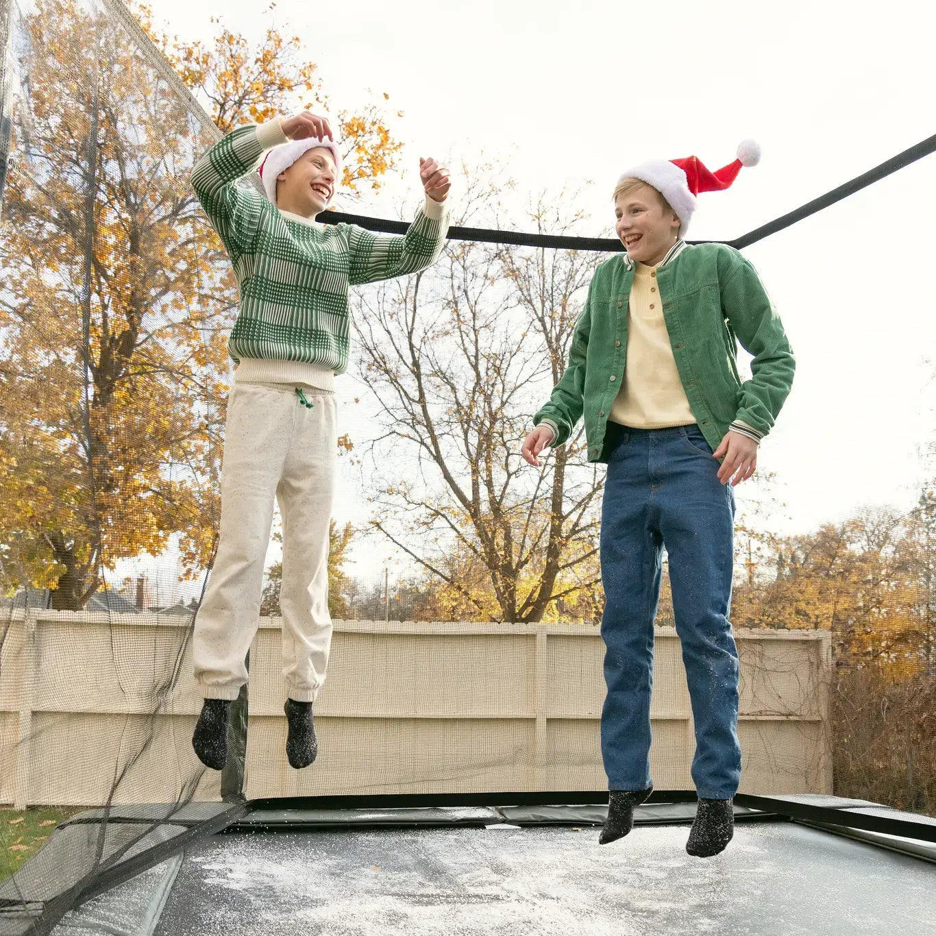 Two kids wearing Santa hats laughing and jumping on a trampoline with light snow.
