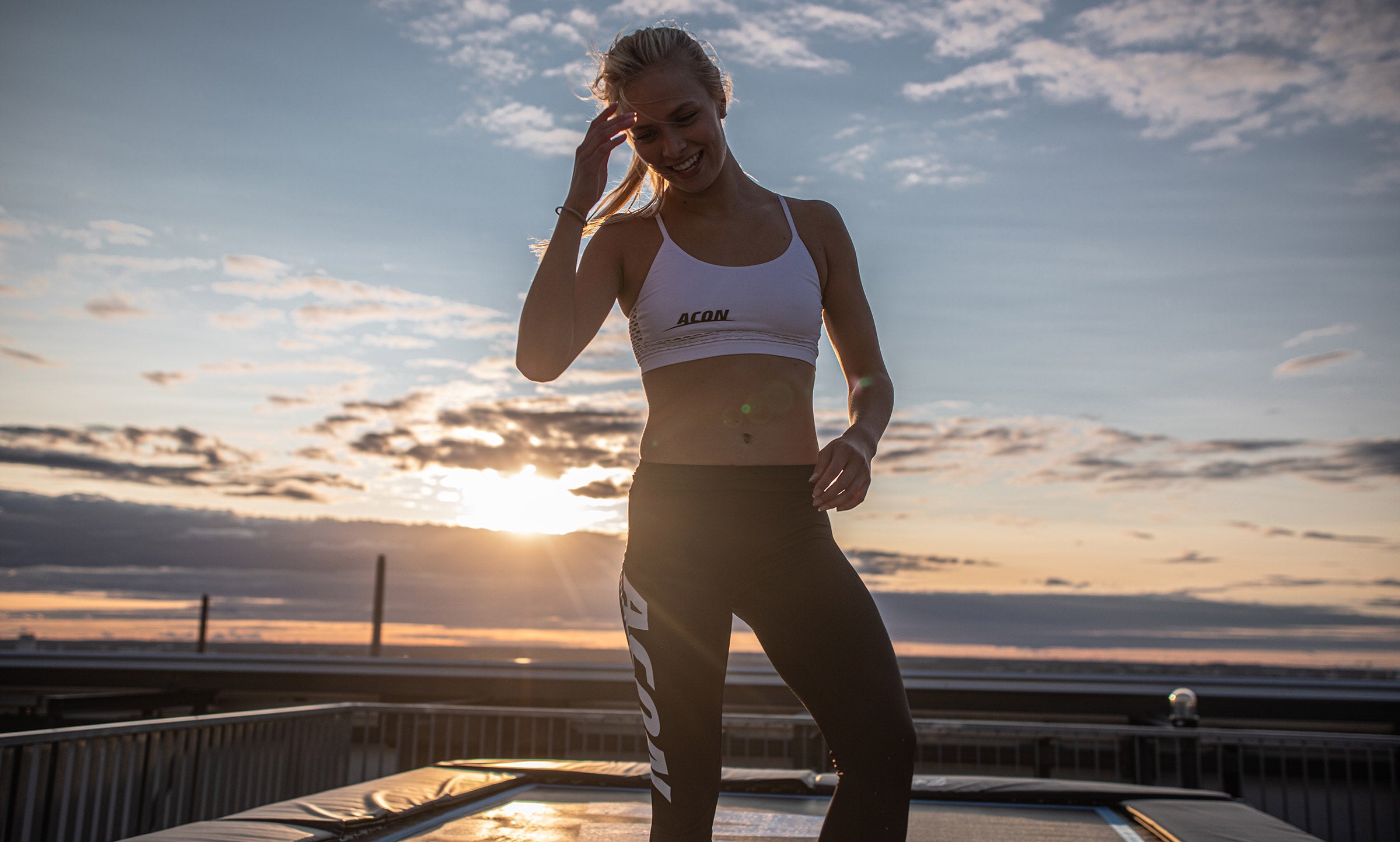 Woman standing on a rectangular trampoline, sunset in the background.