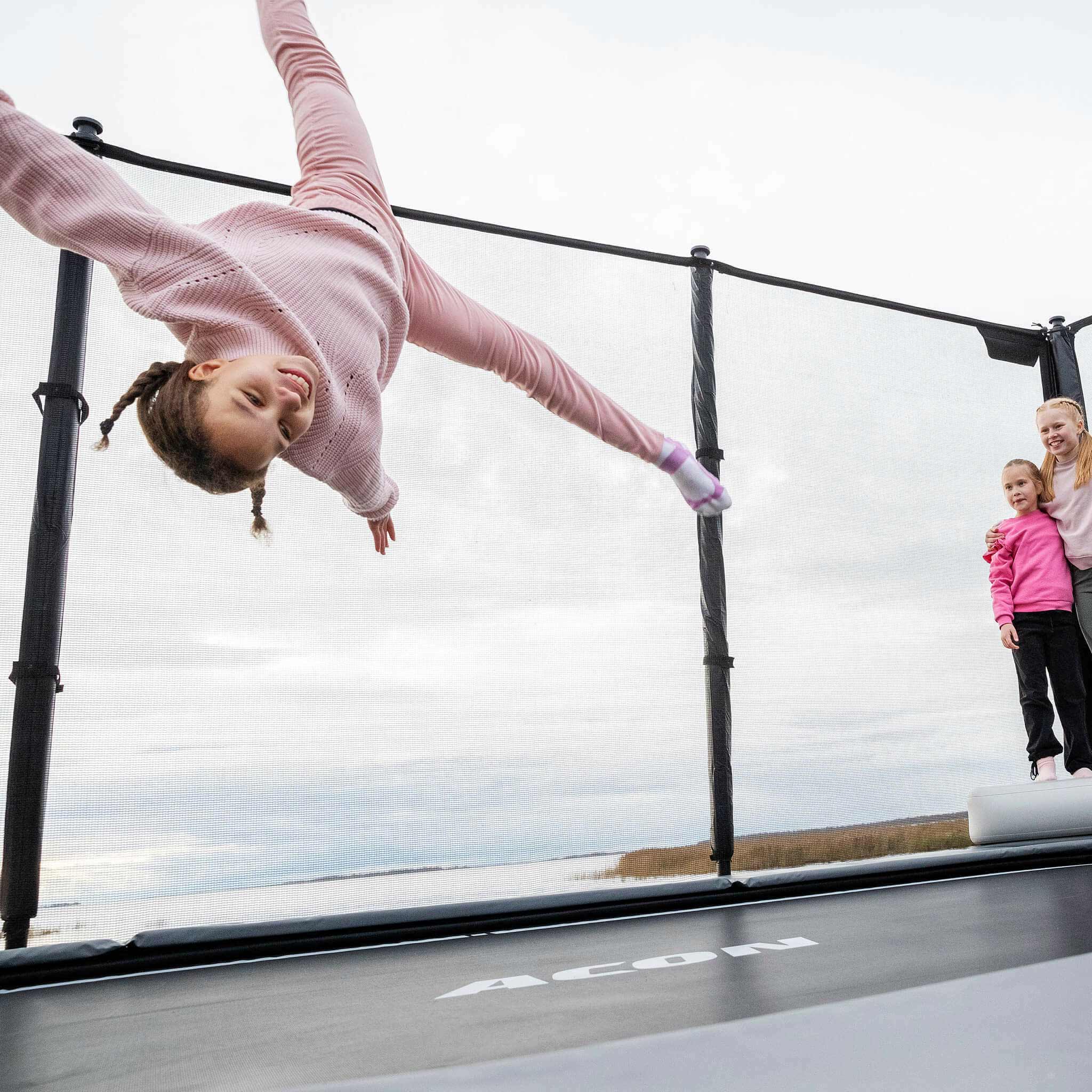 A girl jumps on the Acon X Trampoline, two girls stand and watch.